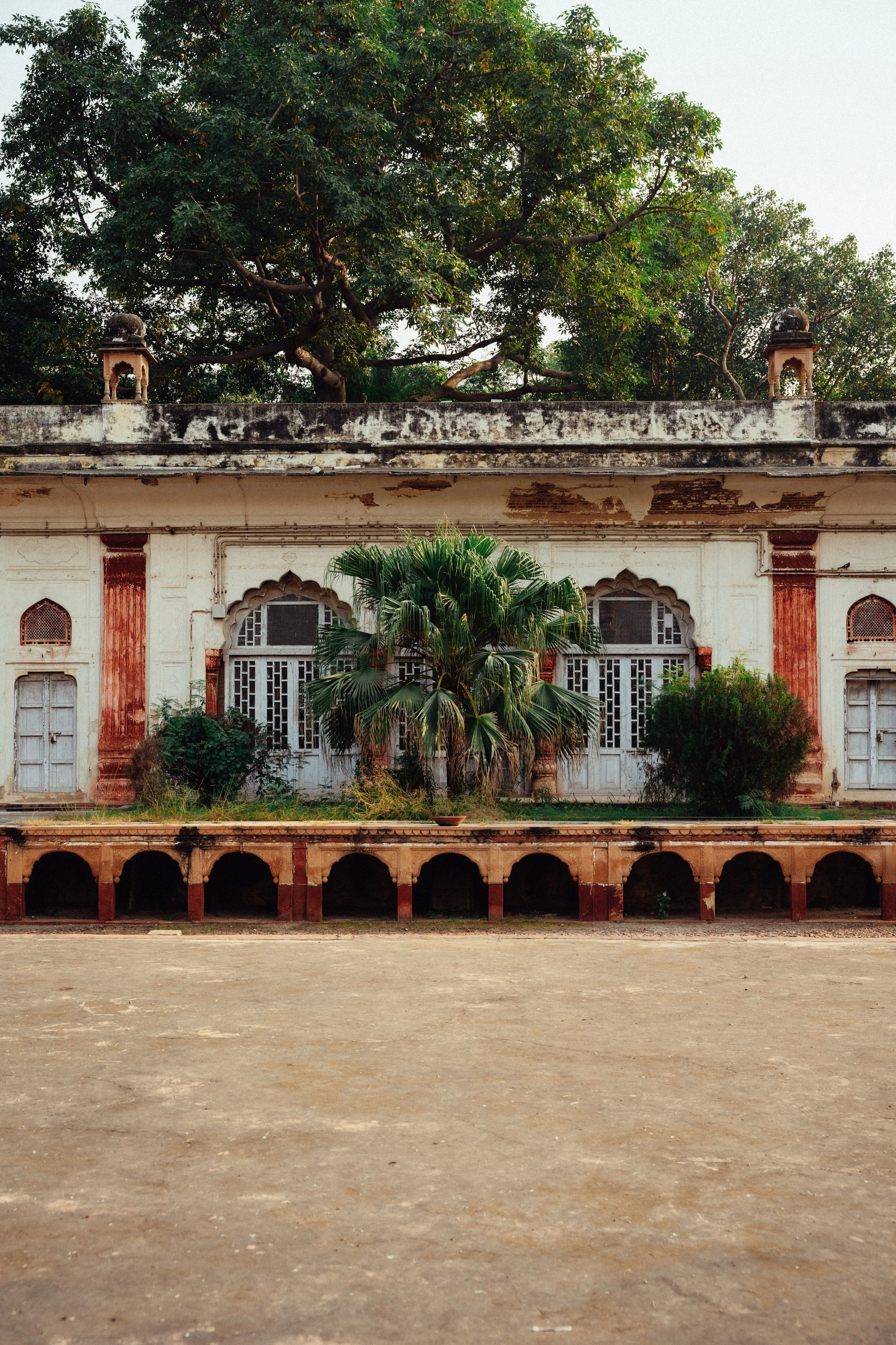 Alter Gebäudeteil mit Pflanzen und Bäumen im Hintergrund, verfallene weiße Wände, offene Arkaden im Vordergrund.