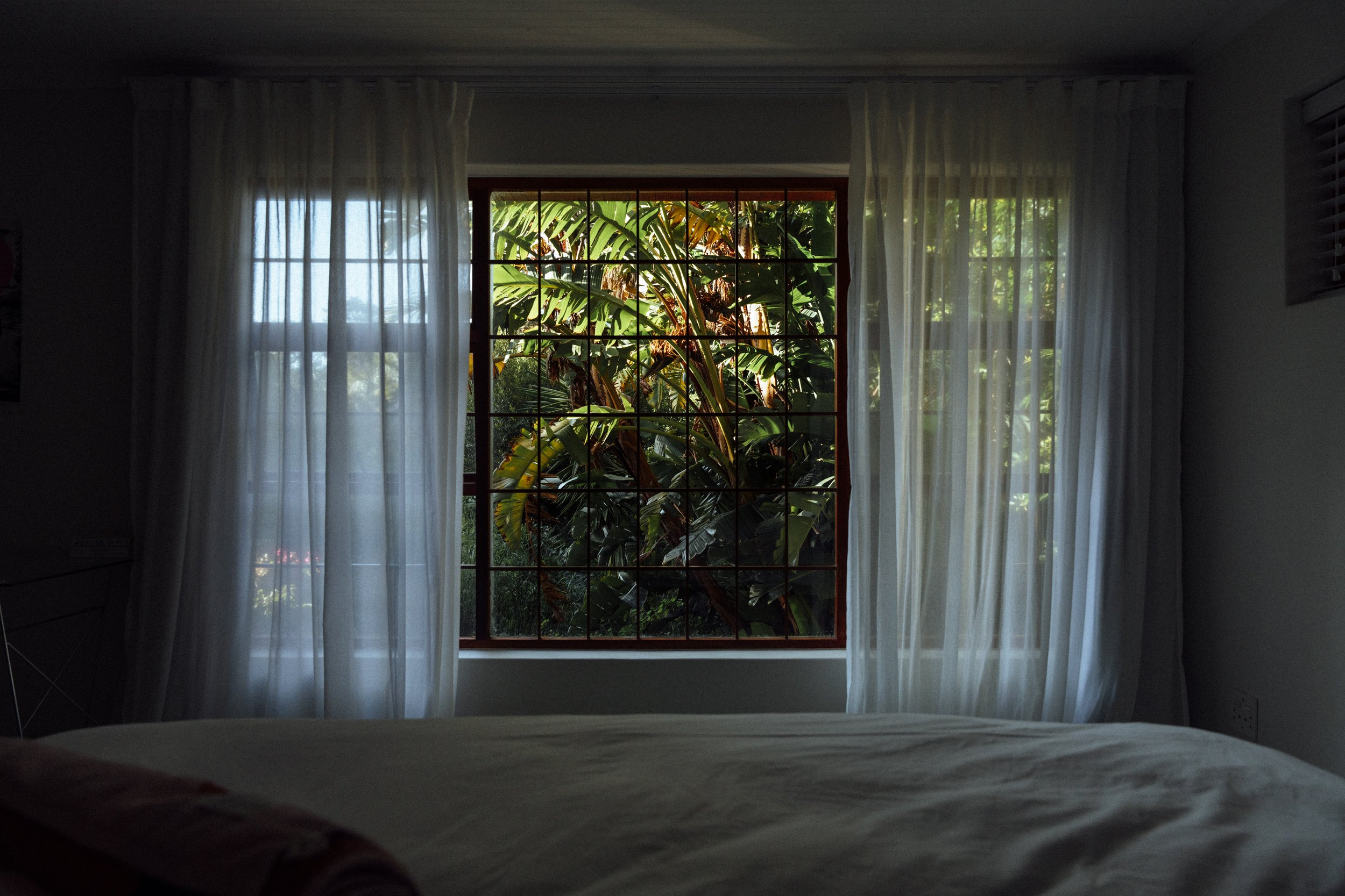 Blick aus einem Schlafzimmer auf eine große Fensterfront mit weißen Gardinen, durch die ein üppiger grüner Garten sichtbar ist.