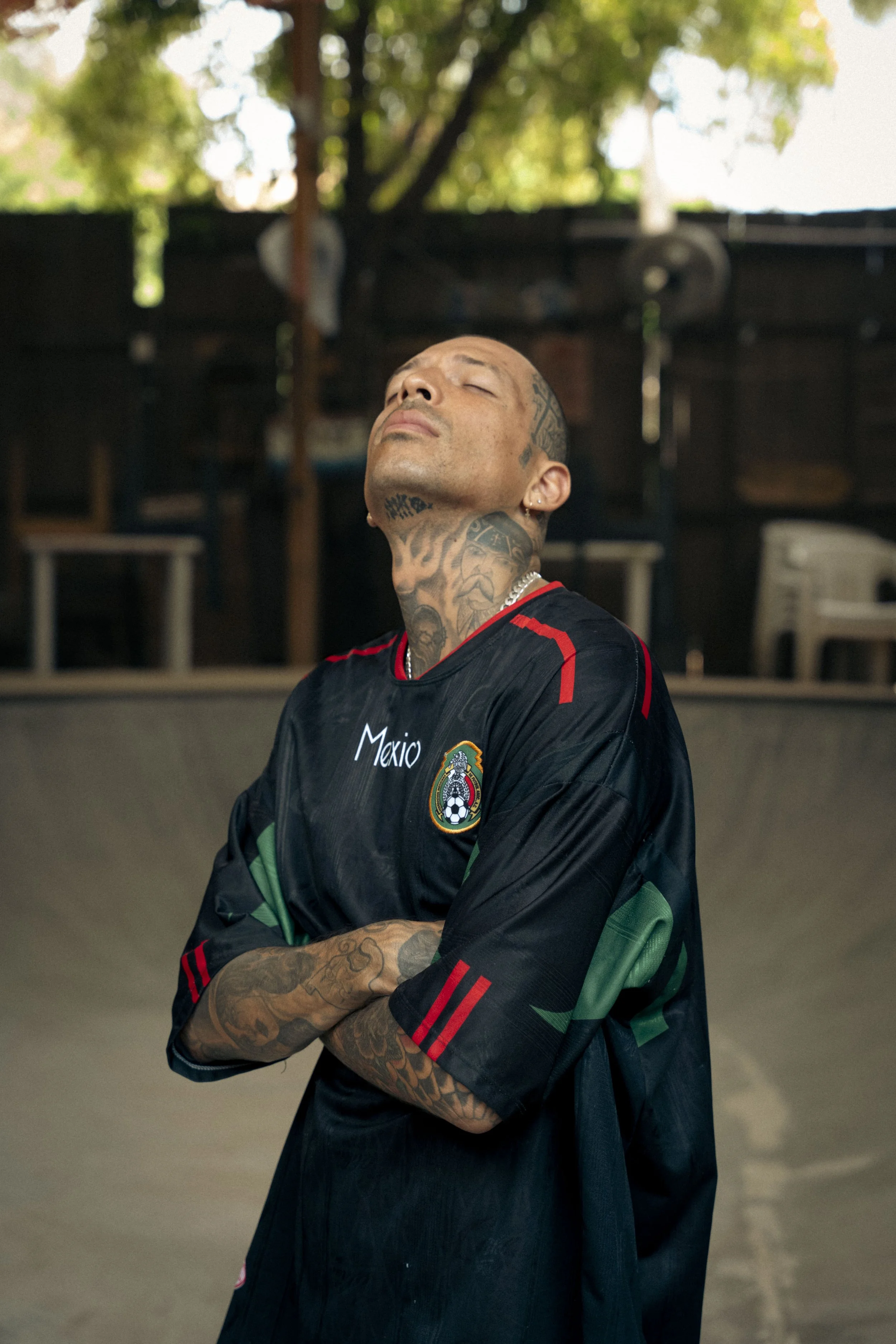 Ein tattoosierter Mann trägt ein mexikanisches Fußballtrikot und steht in einem Skatepark, mit verschränkten Armen, seine Augen sind geschlossen, in einer nachdenklichen Pose.