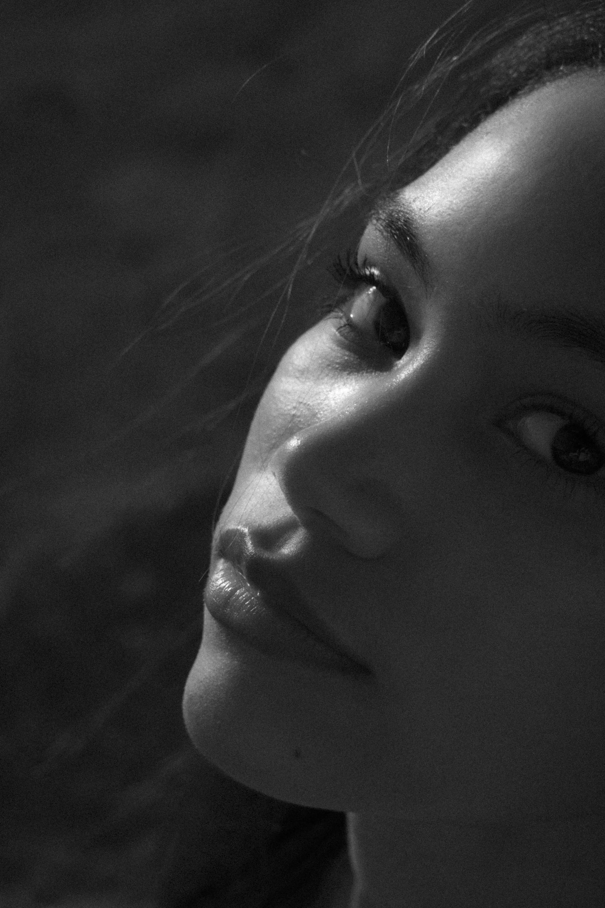 Nahaufnahme eines nachdenklichen Gesichts einer jungen Frau in Schwarz-Weiß, Blick nach oben, weicher Lichtakzent auf Gesicht, dunkler Hintergrund.