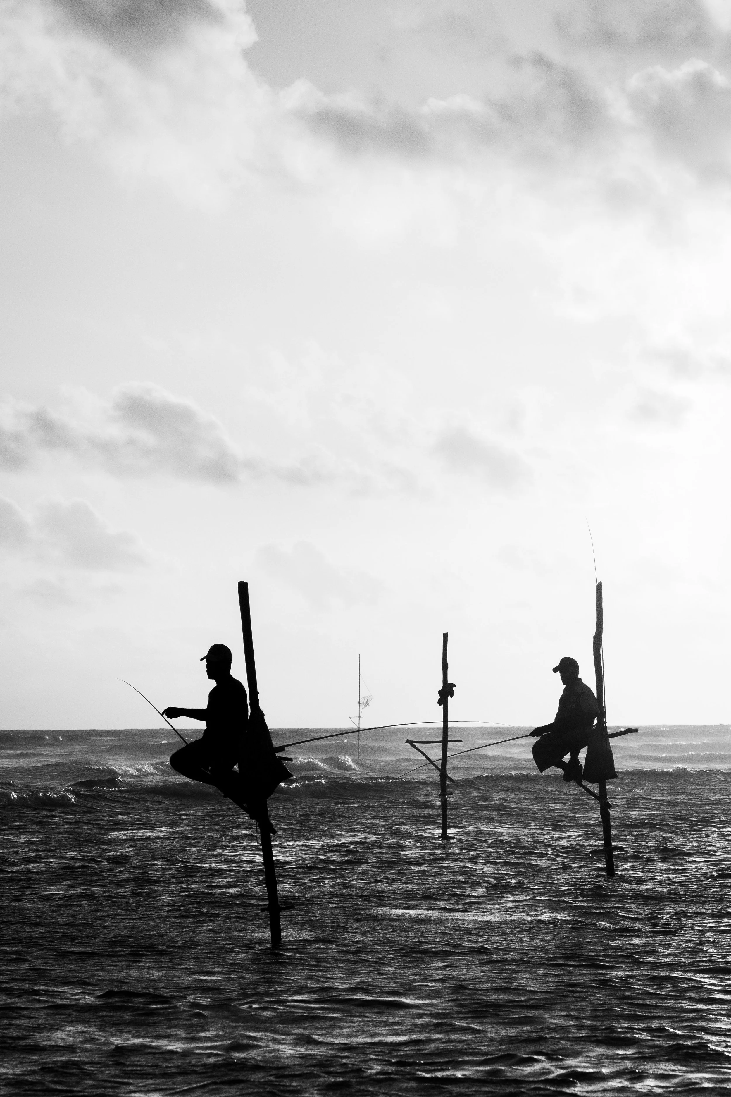 Zwei Menschen, die auf Stangen im Wasser sitzen und mit Angelruten angeln, bei Sonnenuntergang oder -aufgang, schwarz-weiß