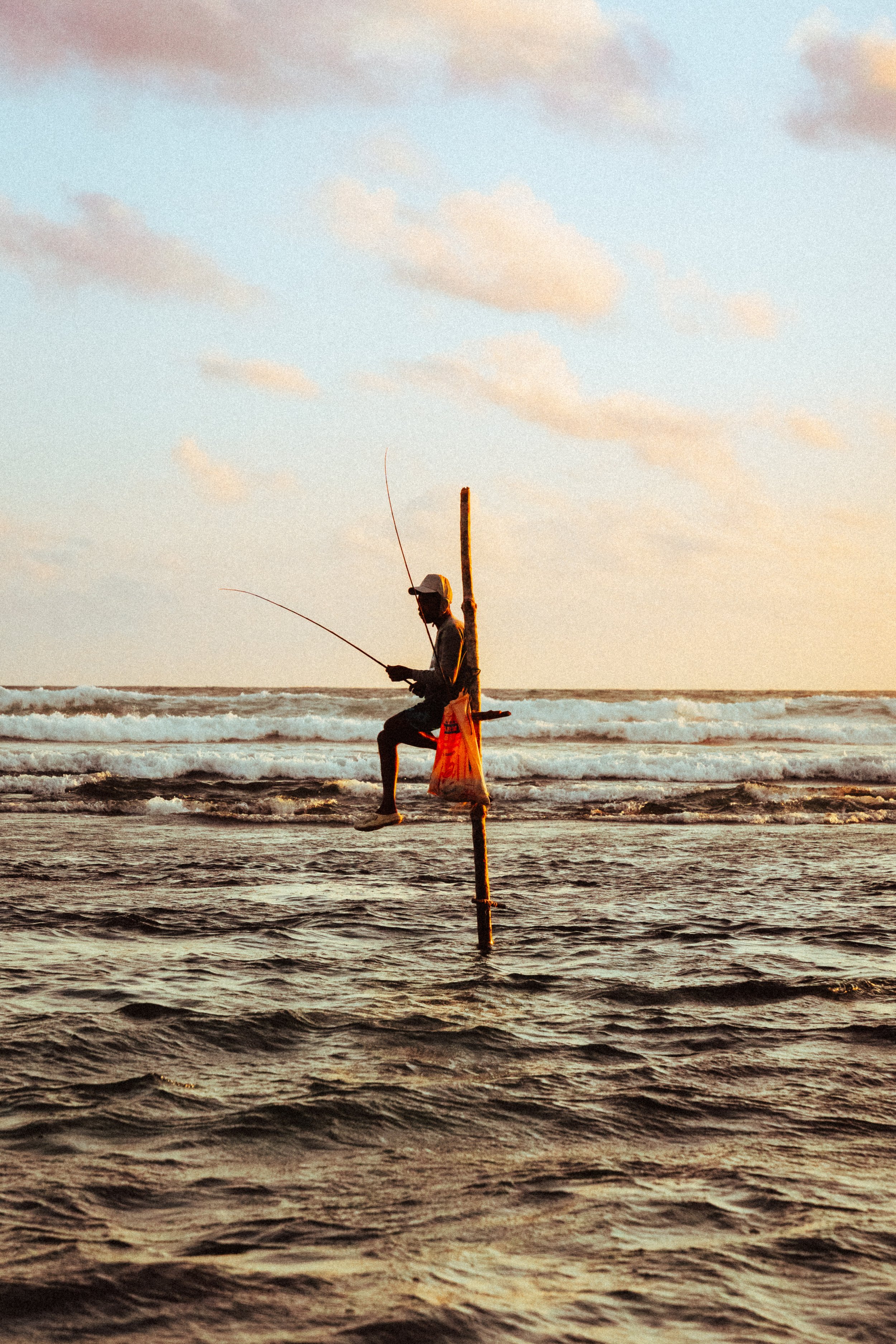 Ein Fischer sitzt auf einem vertikal im Wasser stehendem Holzpfahl und fischt im Meer bei Sonnenuntergang.