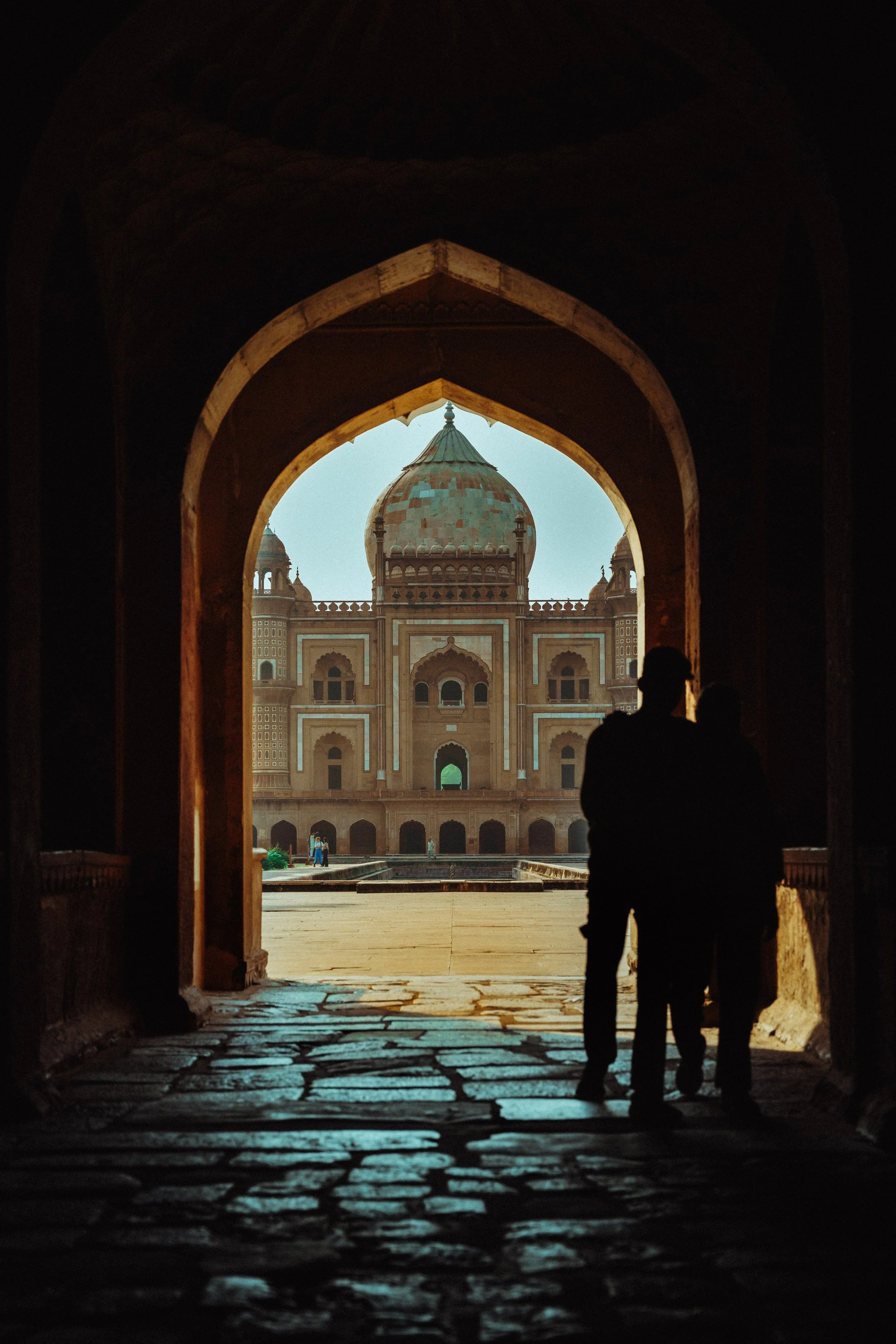 Silhouette eines Paares, das durch einen historischen Torbogen blickt auf ein großes, ehemaliges Palastgebäude mit Kuppeln in Agra, Indien.