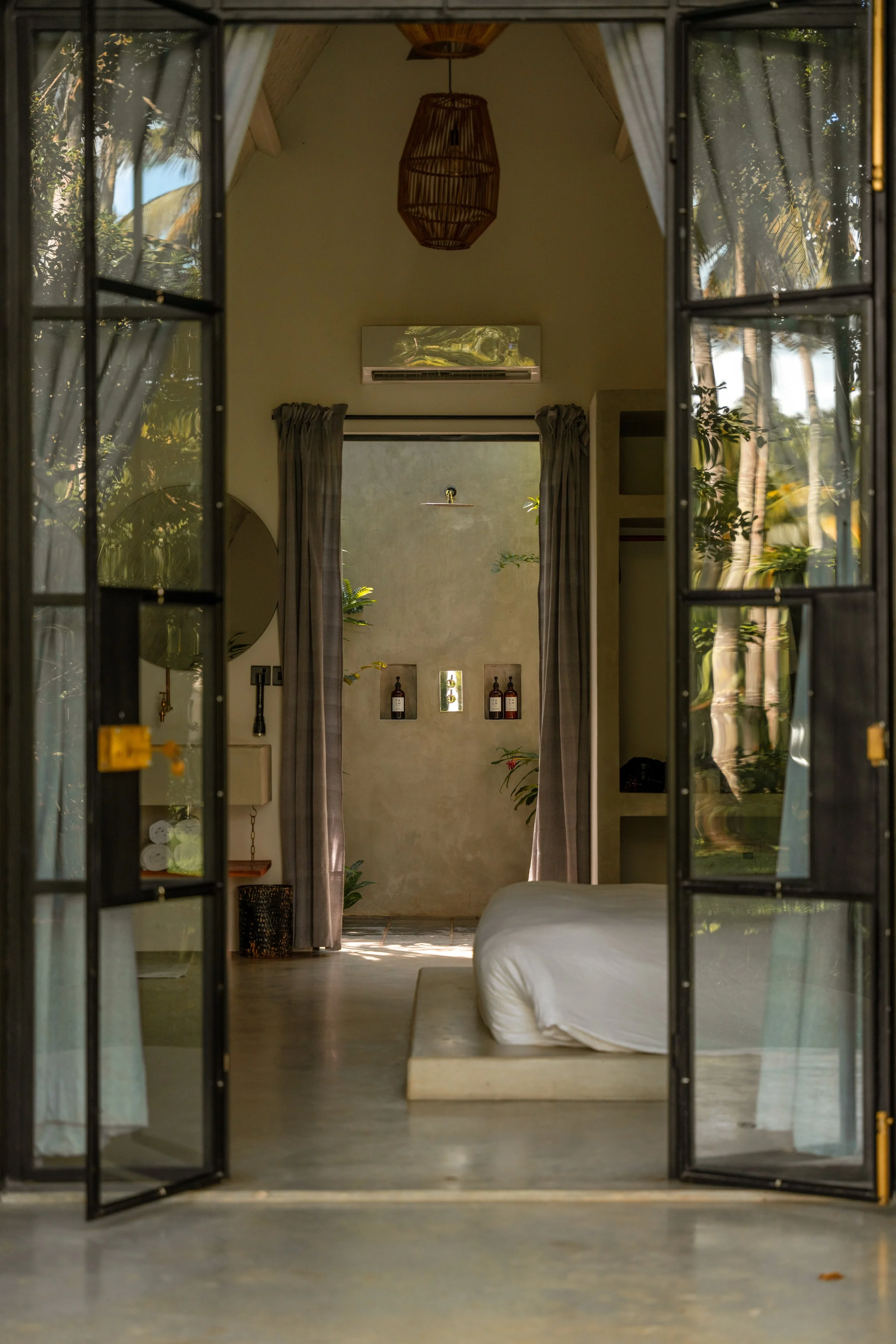 Ein Blick durch offene Glastüren in ein modernes, minimalistisches Schlafzimmer mit einem niedrigen Bett, Vorhängen, Pflanzen und dekorativen Flaschen an der Wand in einer tropischen Umgebung.