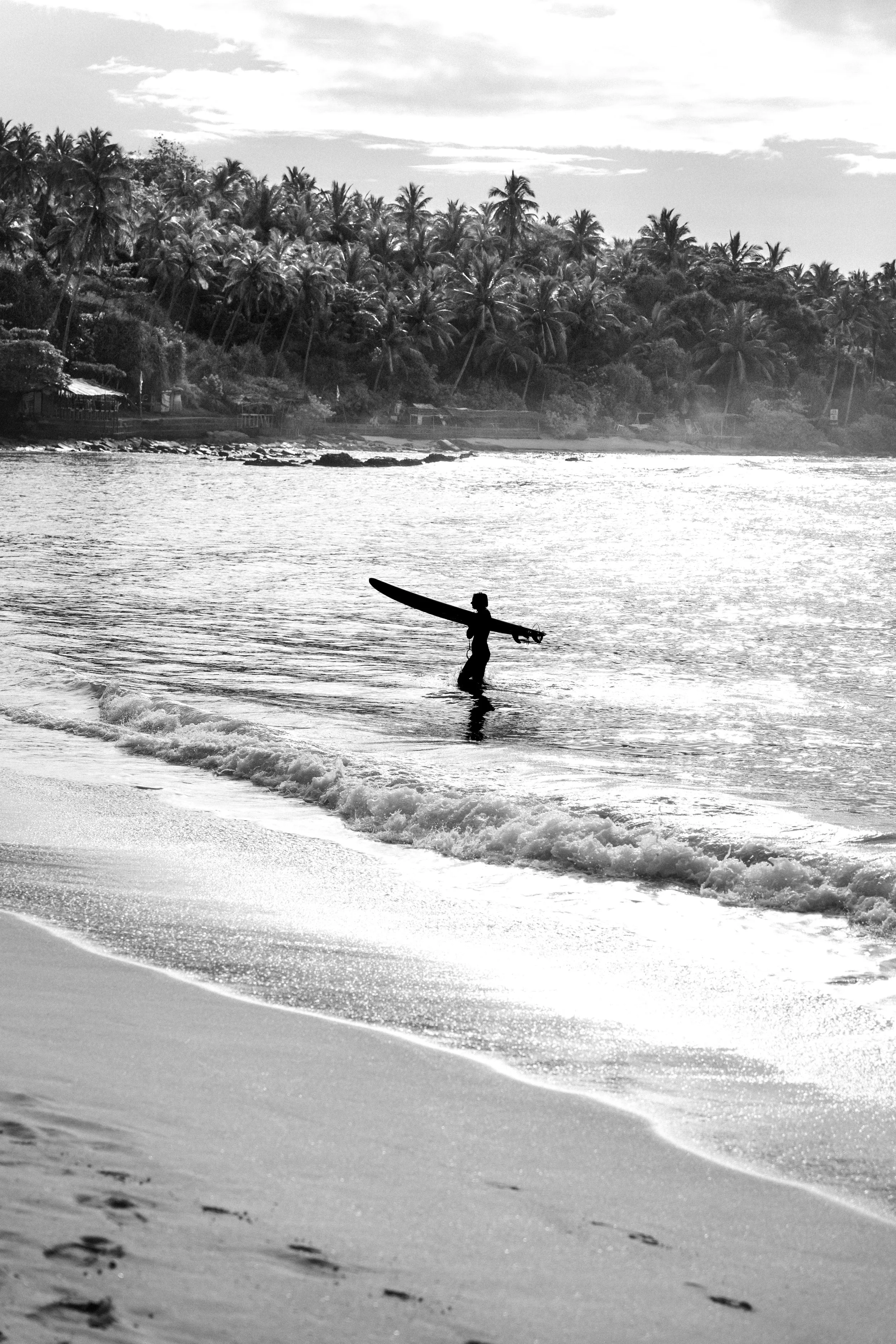 Schwarz-Weiß-Foto eines einzelnen Surfers mit Surfbrett im Wasser an einem Strand, mit Palmen im Hintergrund.