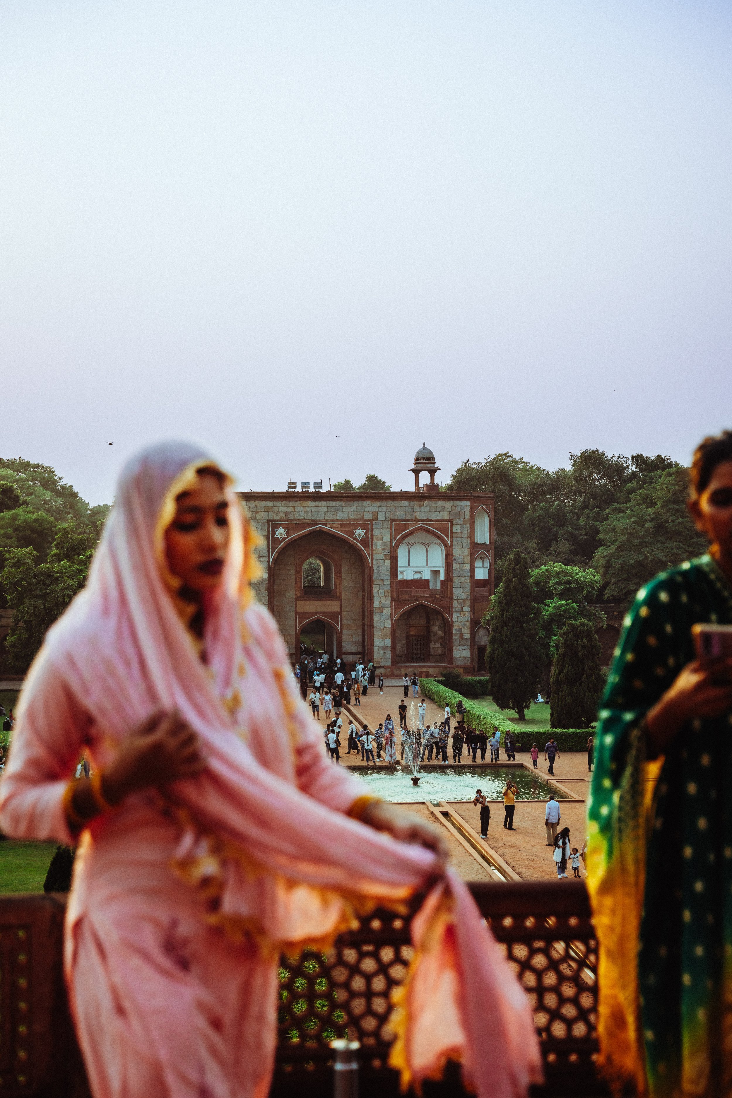 Historischer Ort mit Menschen, einem großen Portal, Wasserbrunnen und grünen Bäumen im Hintergrund, einige Menschen im Vordergrund in traditioneller Kleidung.