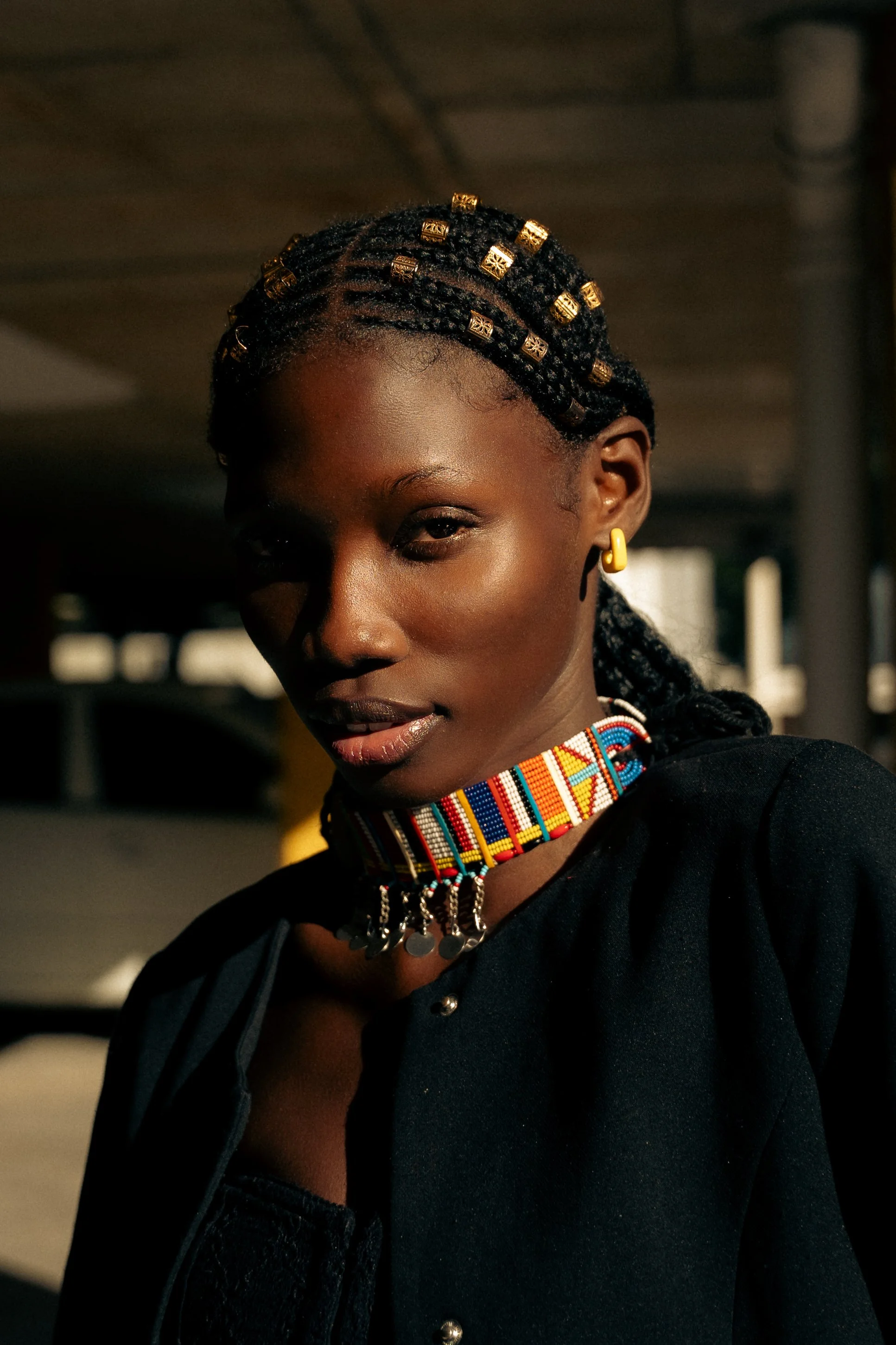 Porträt einer schwarzen Frau mit geflochtenem Haar, Gold-Accessoires, buntem Perlenhalsband, schwarze Kleidung, im Schatten bei Sonnenlicht aufgenommen.