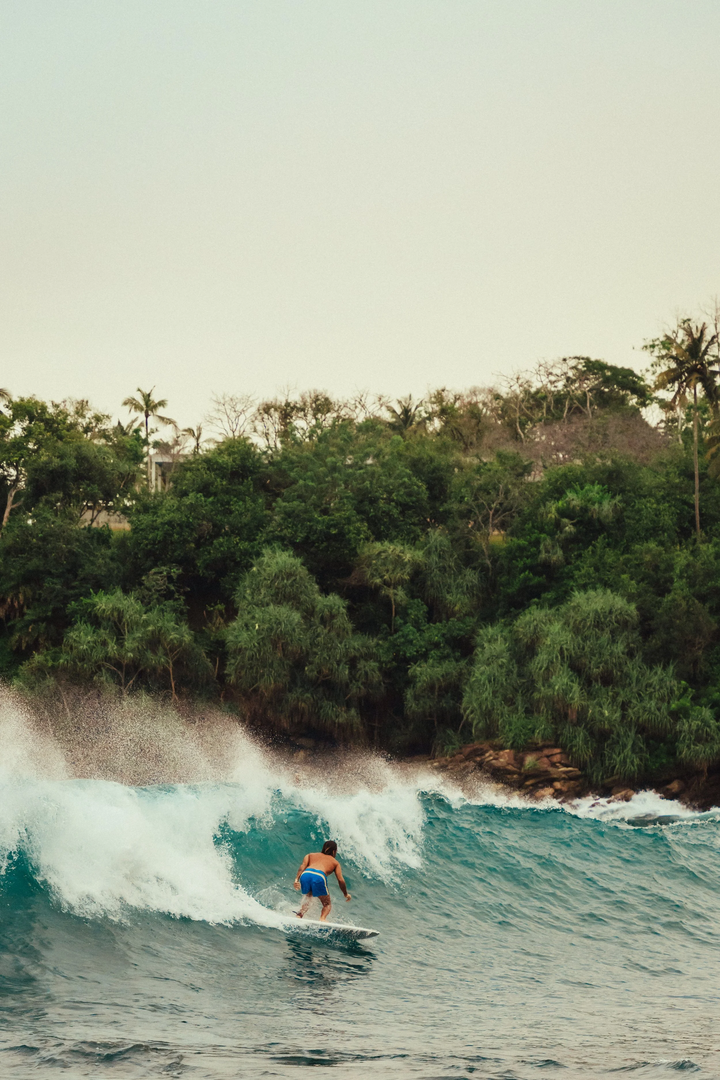 Ein Mann surft auf einem Meer mit Wellen in einer Bucht, umgeben von grünen, bewaldeten Hügeln.