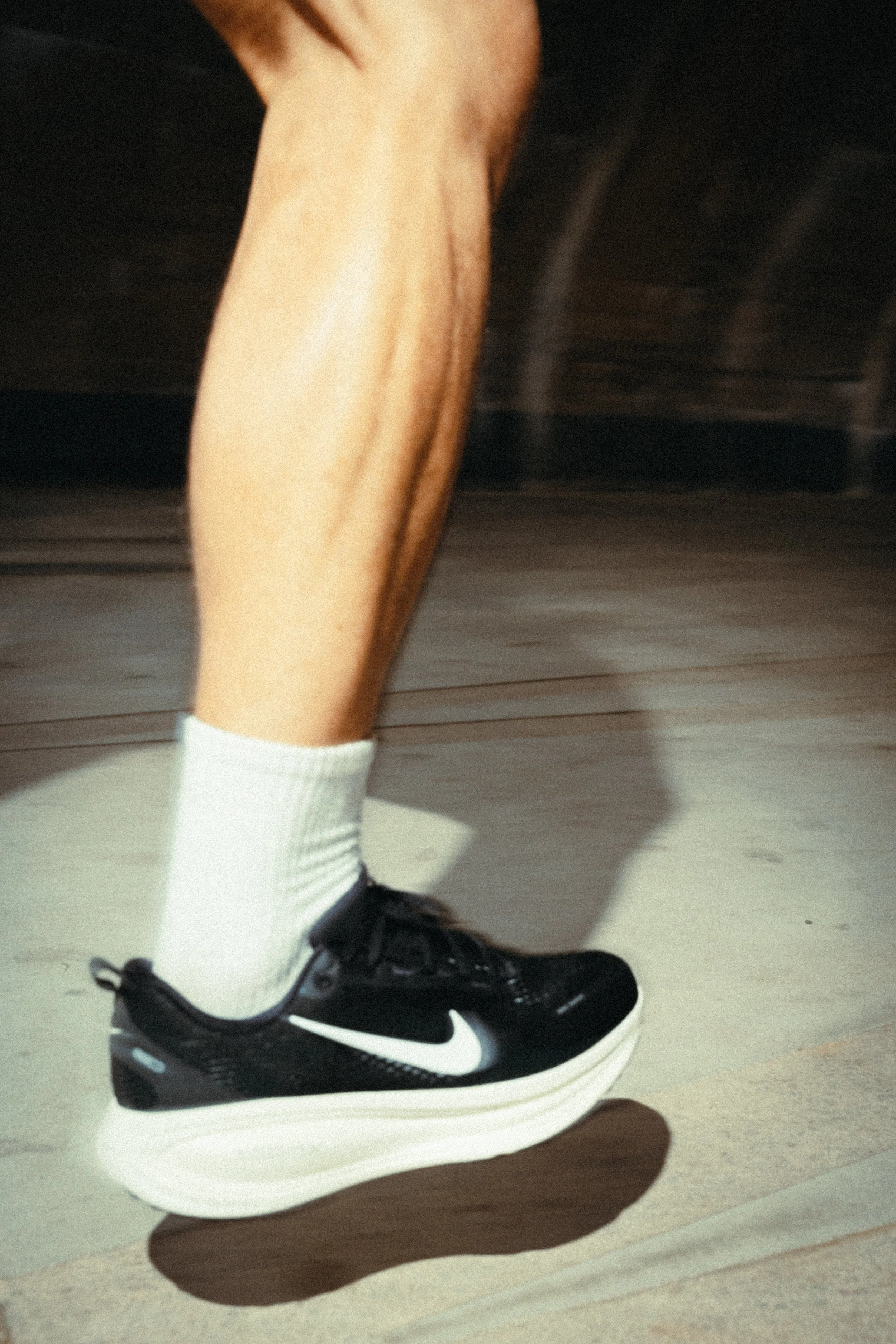 Ein Bein mit weißem Socken, schwarzem Nike-Laufschuh auf glattem Boden im Innenraum.