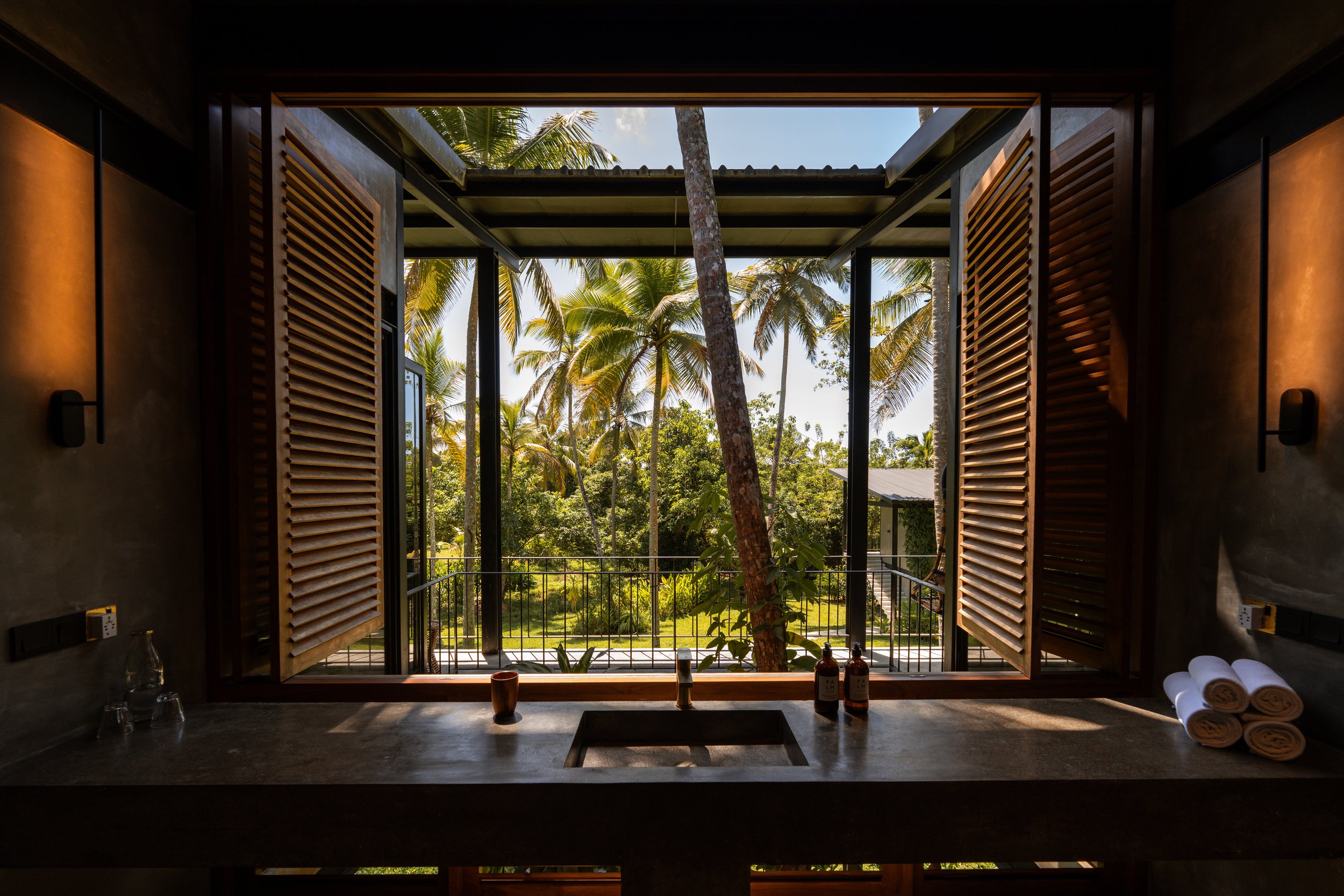 Blick aus einem Badezimmerfenster mit Holzbalkonen auf einen Garten mit Palmen.
