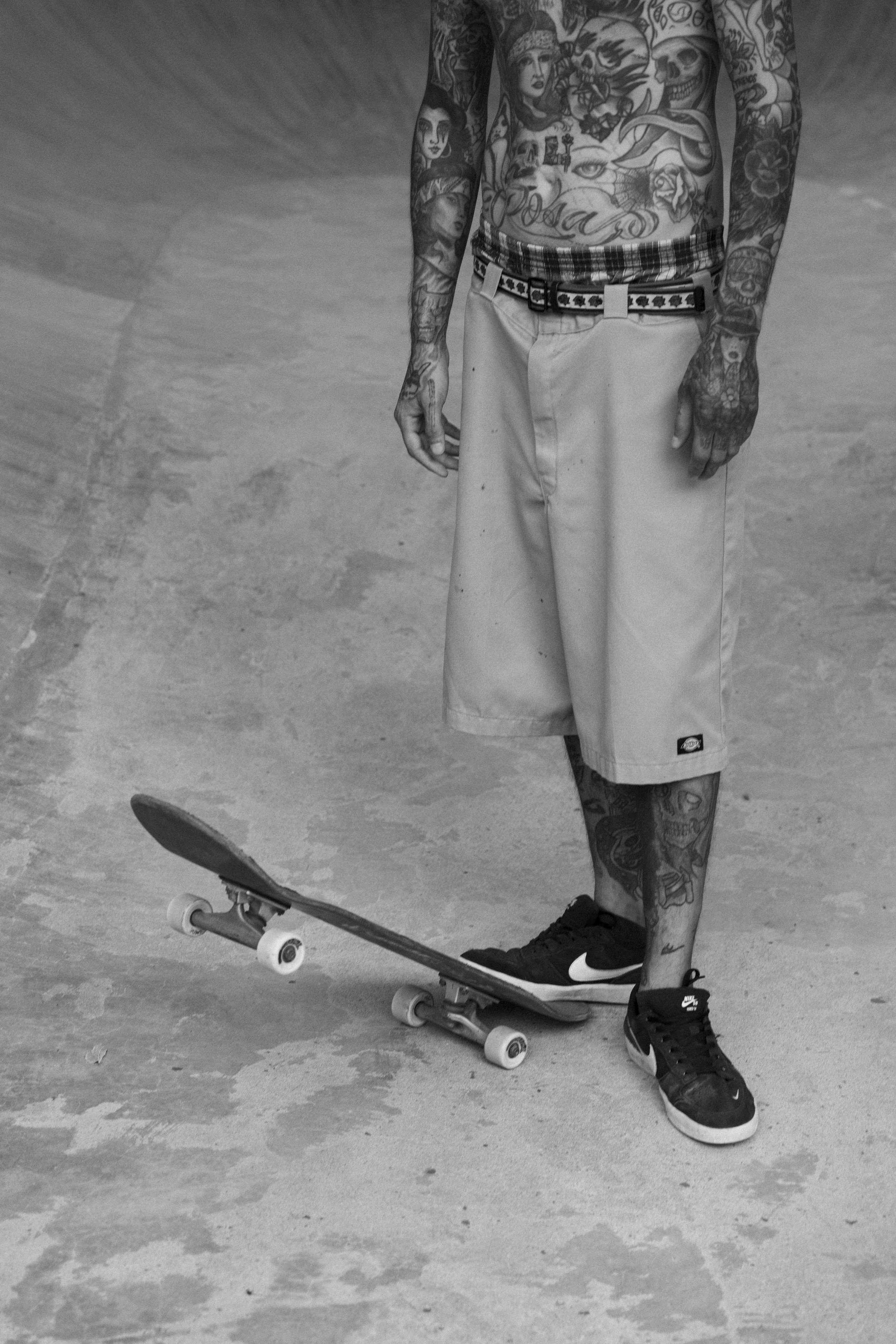 Ein tattoo-übersäter Mann trägt Shorts und Nike-Sneaker, steht auf einem Skateboard im Skatepark.
