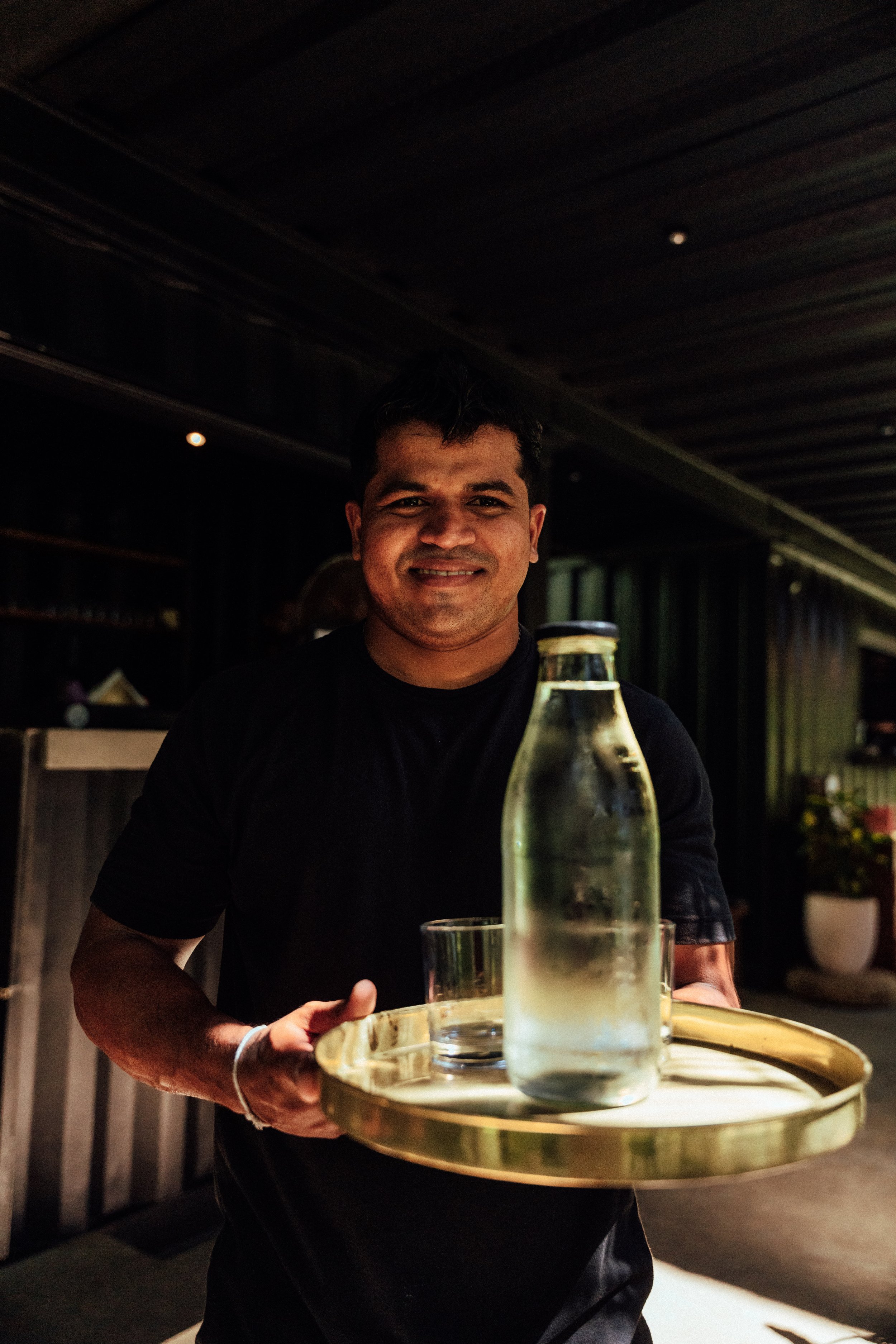 Ein Mann serviert Wasser in einer Bar, hält einen runden goldfarbenen Tablett mit Wasserflasche und Glas.