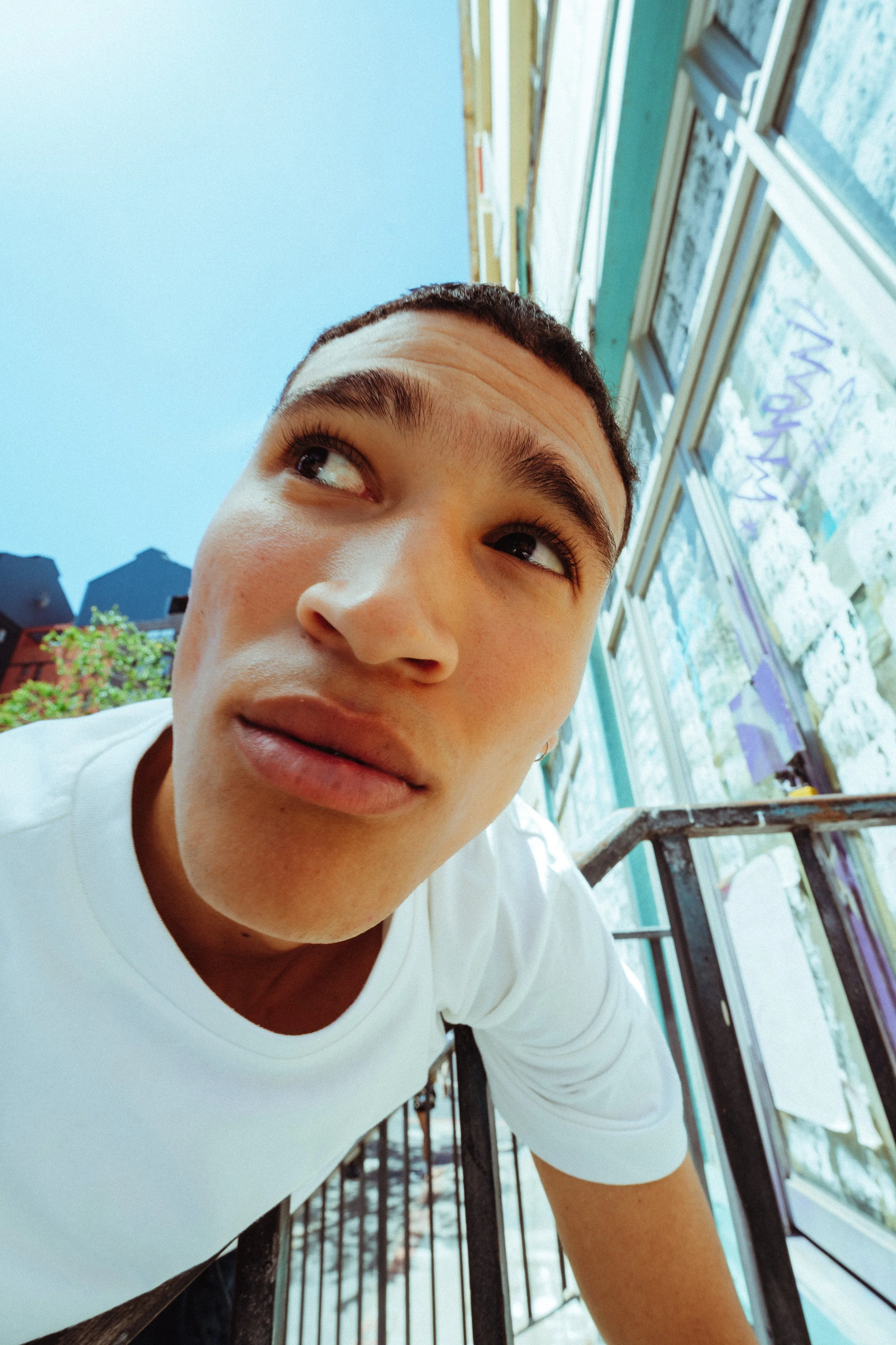 Nahaufnahme eines jungen Mannes, der auf einem Balkon steht, mit Blick nach oben, gegen einen blauen Himmel, im Hintergrund Häuser und Bäume.