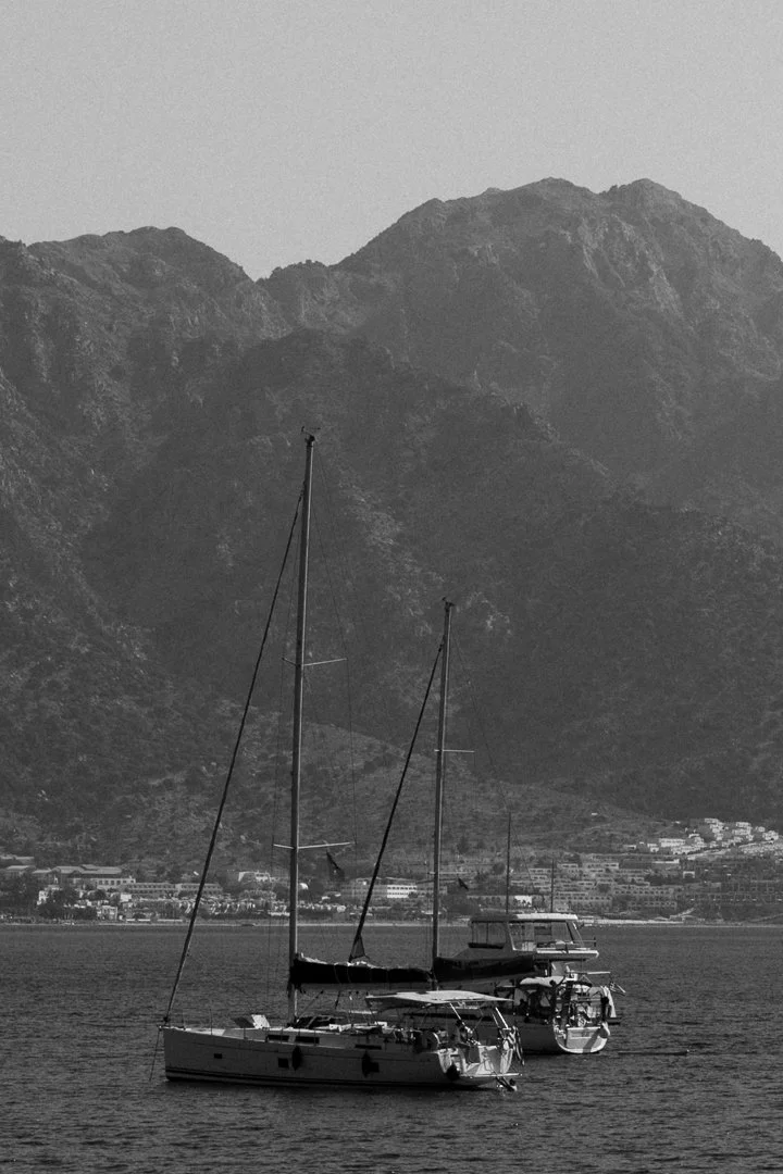 Zwei Segelboote auf einem ruhigen See mit Bergkulisse im Hintergrund, schwarz-weiße Aufnahme.