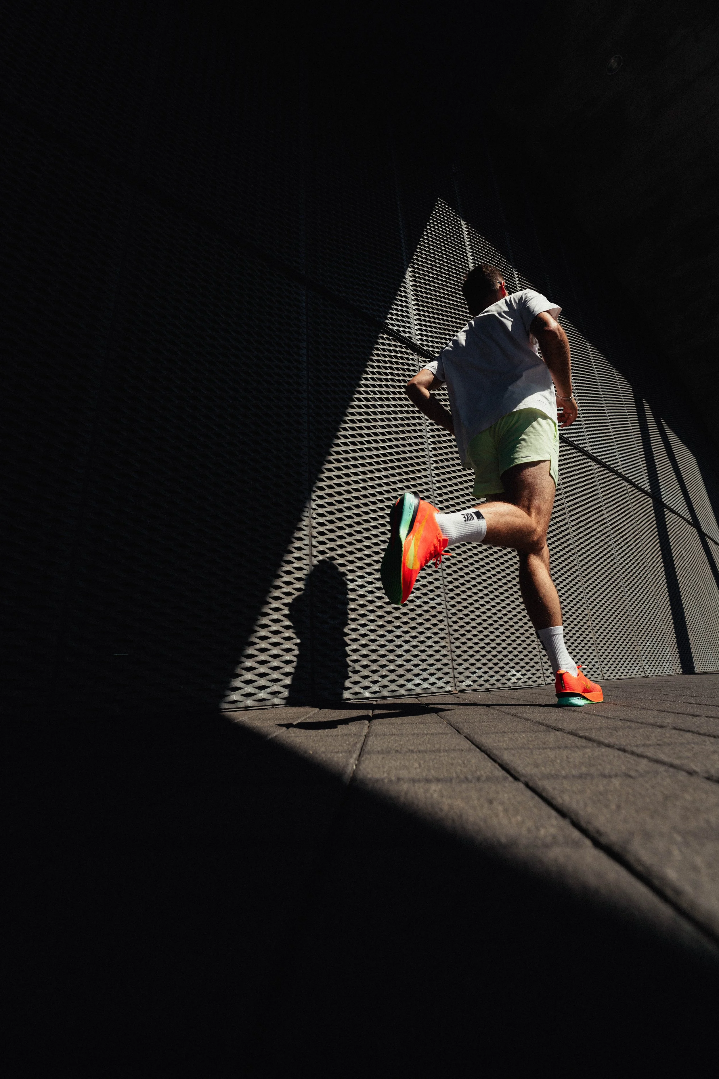 Ein Mann läuft an einer Metallwand vorbei, trägt weiße Sportschuhe mit neonorangen Akzenten, weiße Socken, grüne Shorts und ein graues T-Shirt, bei starker Sonneneinstrahlung, mit Schatten auf dem Boden.
