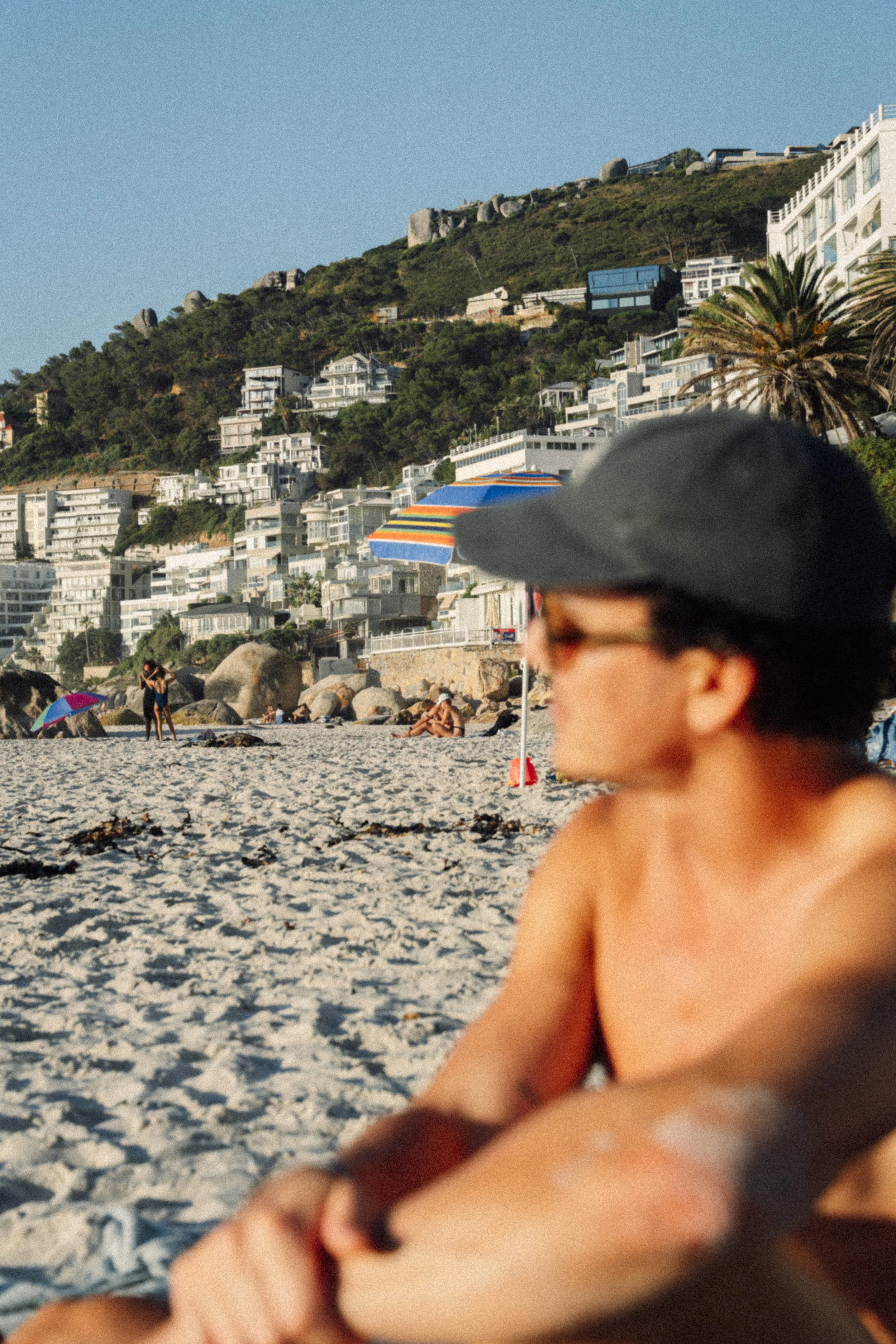 Ein Strand mit Menschen, einem bunten Sonnenschirm und Gebäuden auf einer Hanglage im Hintergrund, eine Person trägt eine Cap und Sonnenbrille.
