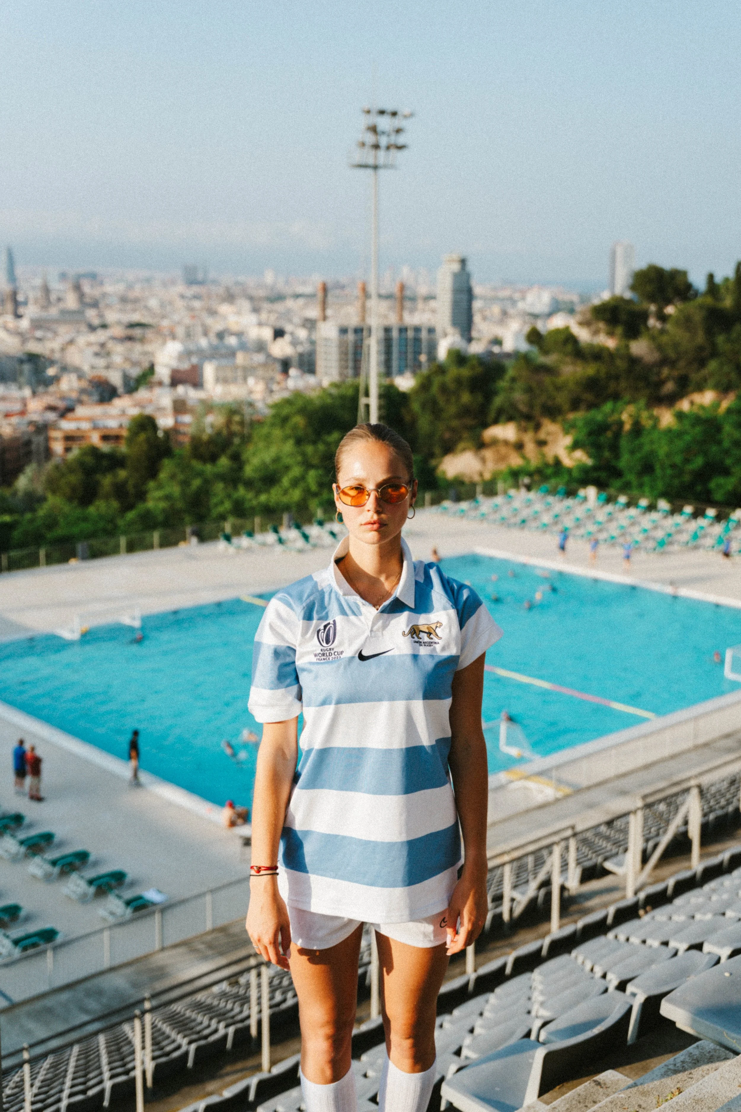Junge Frau in argentinischer Fußballnationalmannschaftsjacke vor einem Pool mit Stadt im Hintergrund.