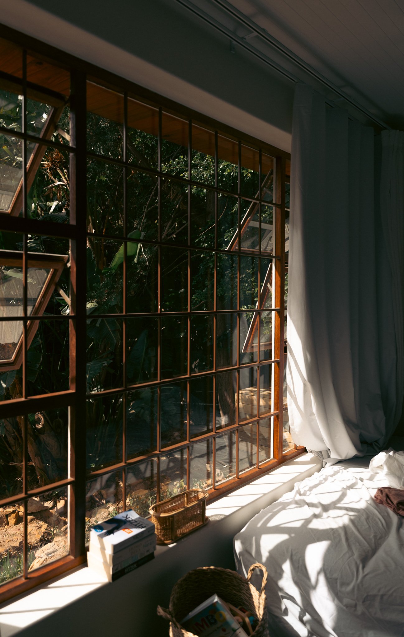 Blick durch ein großes, rechteckiges Fenster mit Holzrahmen auf einen Garten mit Bäumen und Sträuchern. Im Innenraum steht ein ungemachtes Bett mit weißen Laken, an der Bettkante stehen eine kleine Papierbox und eine Kiste mit Zeitschriften, ein Müll