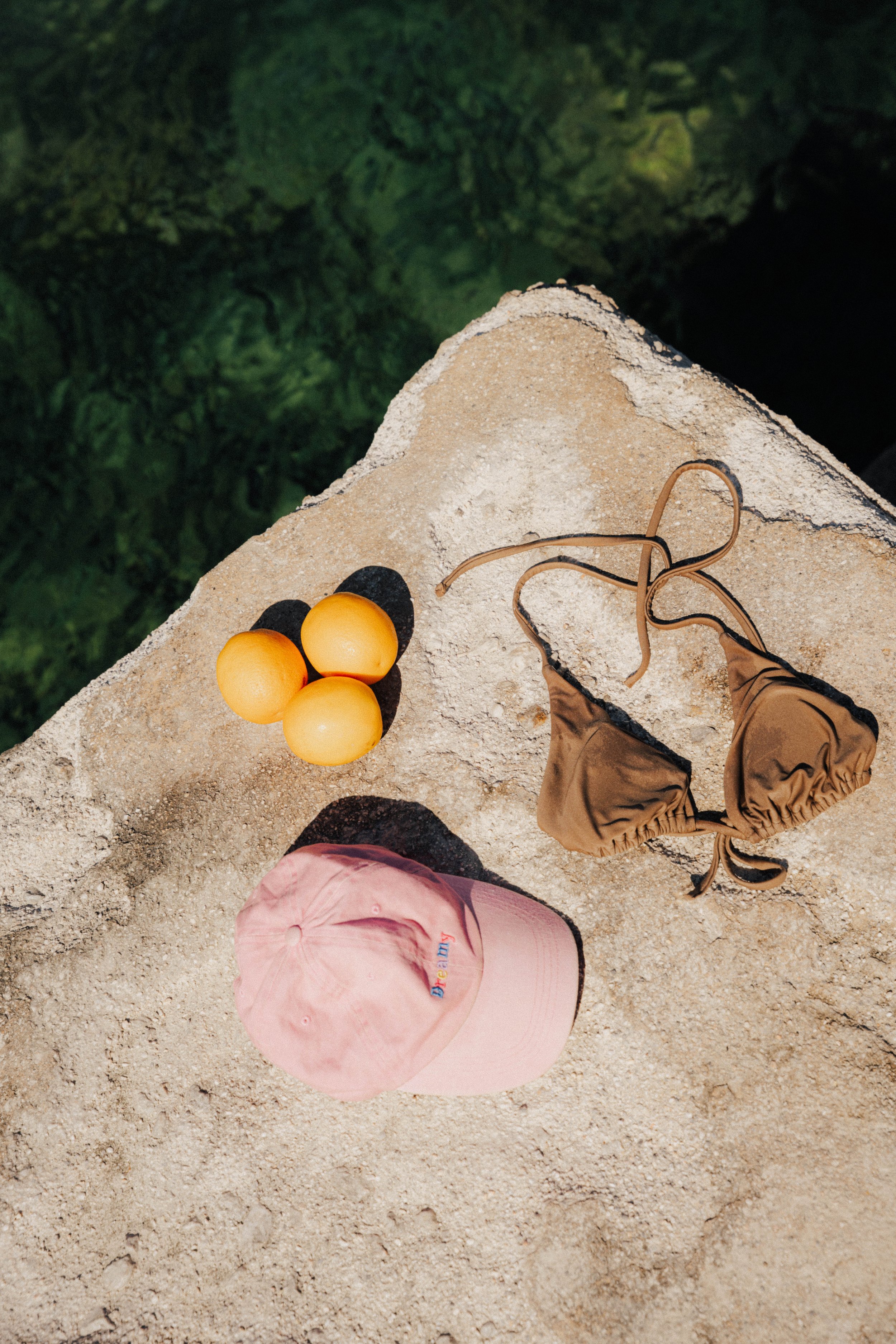 Drei gelbe Pfirsiche, ein rosa Baseballcap, eine braune Bikini-Top auf einer sandigen Oberfläche. Dunkler Wald im Hintergrund.