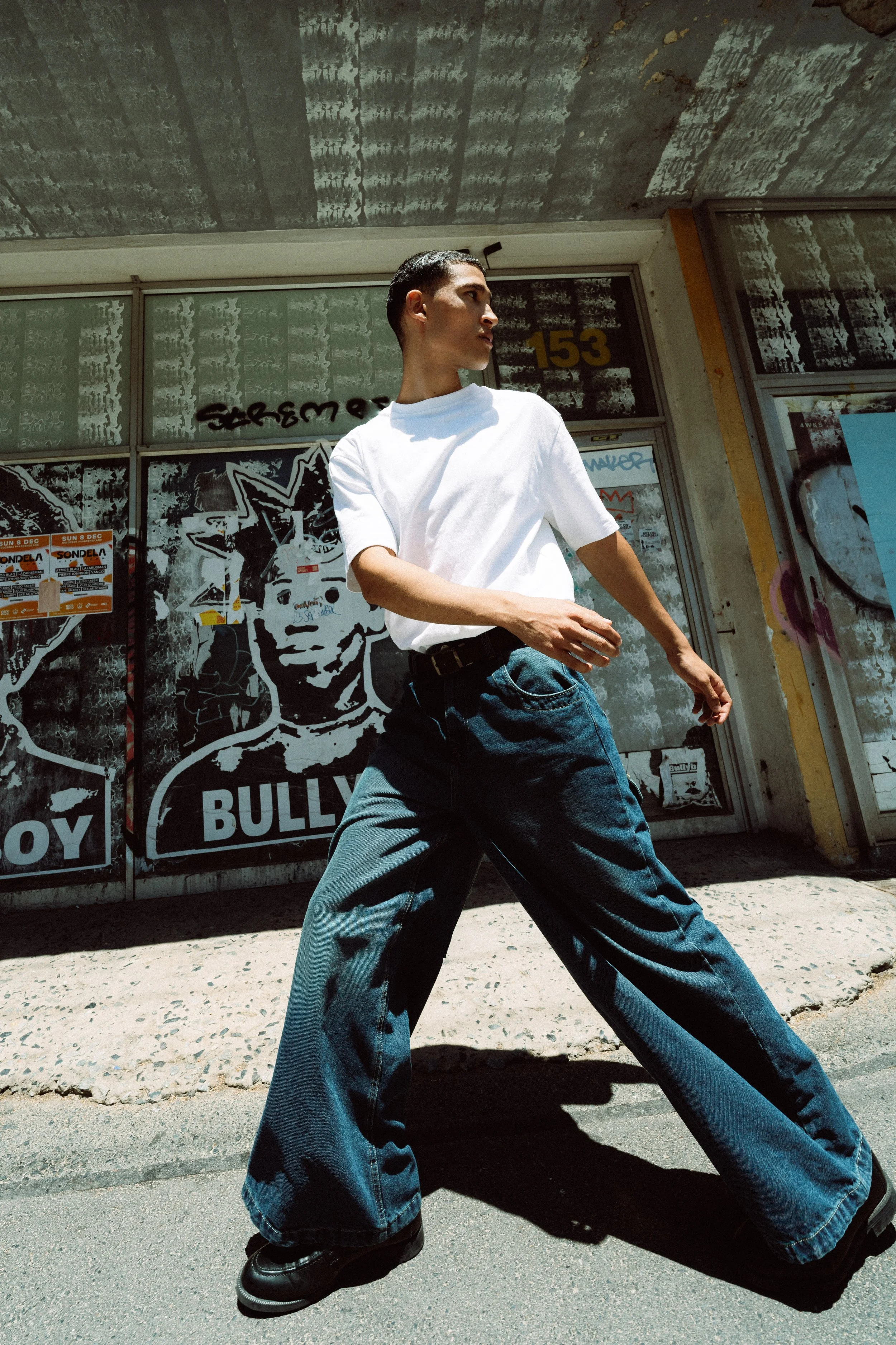 Junger Mann mit weißen T-Shirt und weiten Jeans geht an einem Graffiti-Panel auf der Straße vorbei.