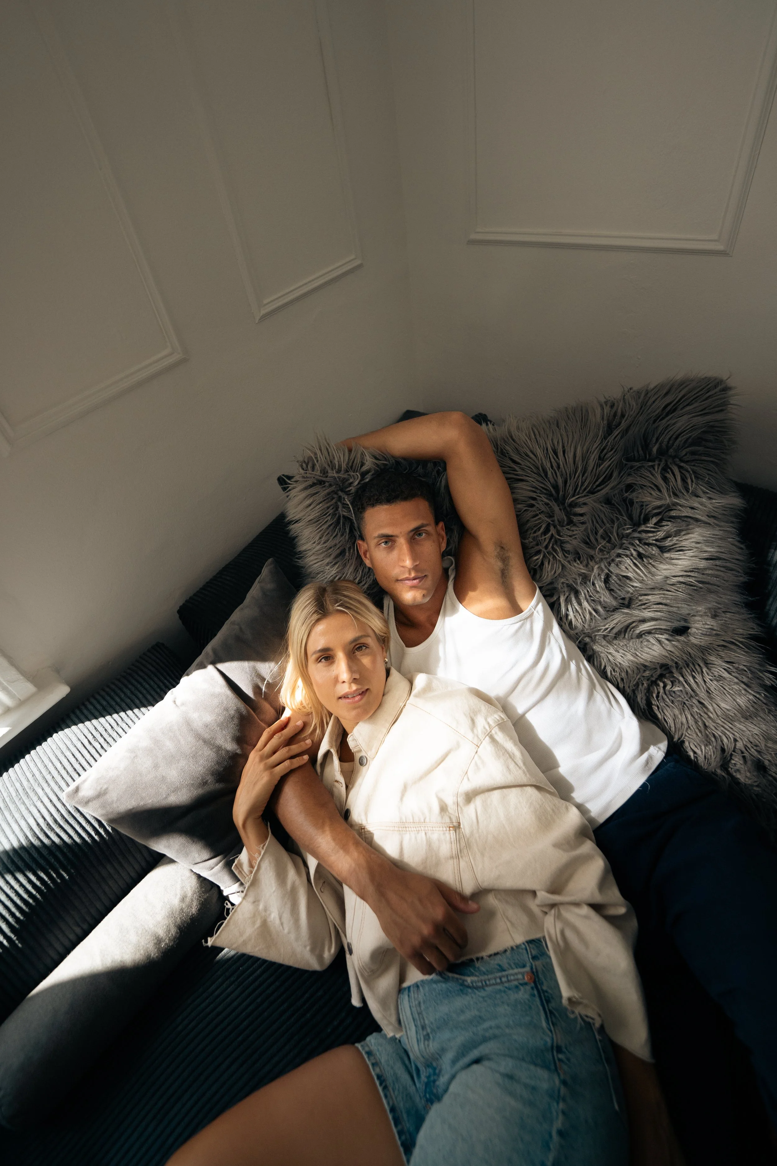 Ein Paar liegt auf einem schwarzen Sofa, umgeben von verschiedenen Kissen, in einem Raum mit weißen Wänden.