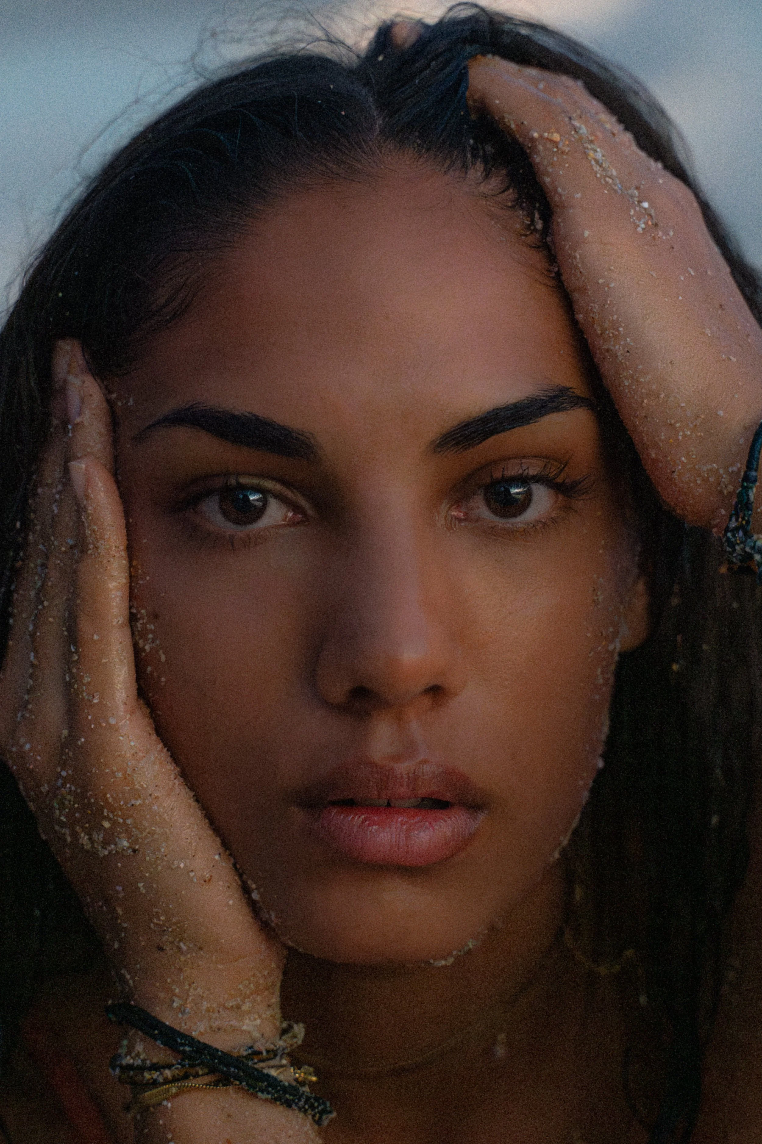 Nahaufnahme eines jungen Mannes mit nassen, sandbedeckten Haaren und Haut, der die Hände an den Kopf legt, Blick direkt in die Kamera, bei Sonnenuntergang