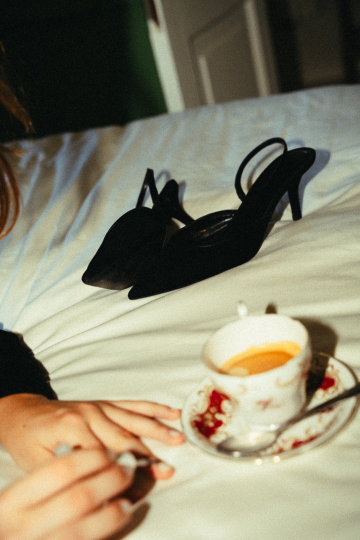 Eine schwarze High Heel-Schuhe, eine Tasse Kaffee auf einem weißen Bett, eine Hand, die etwas hält.