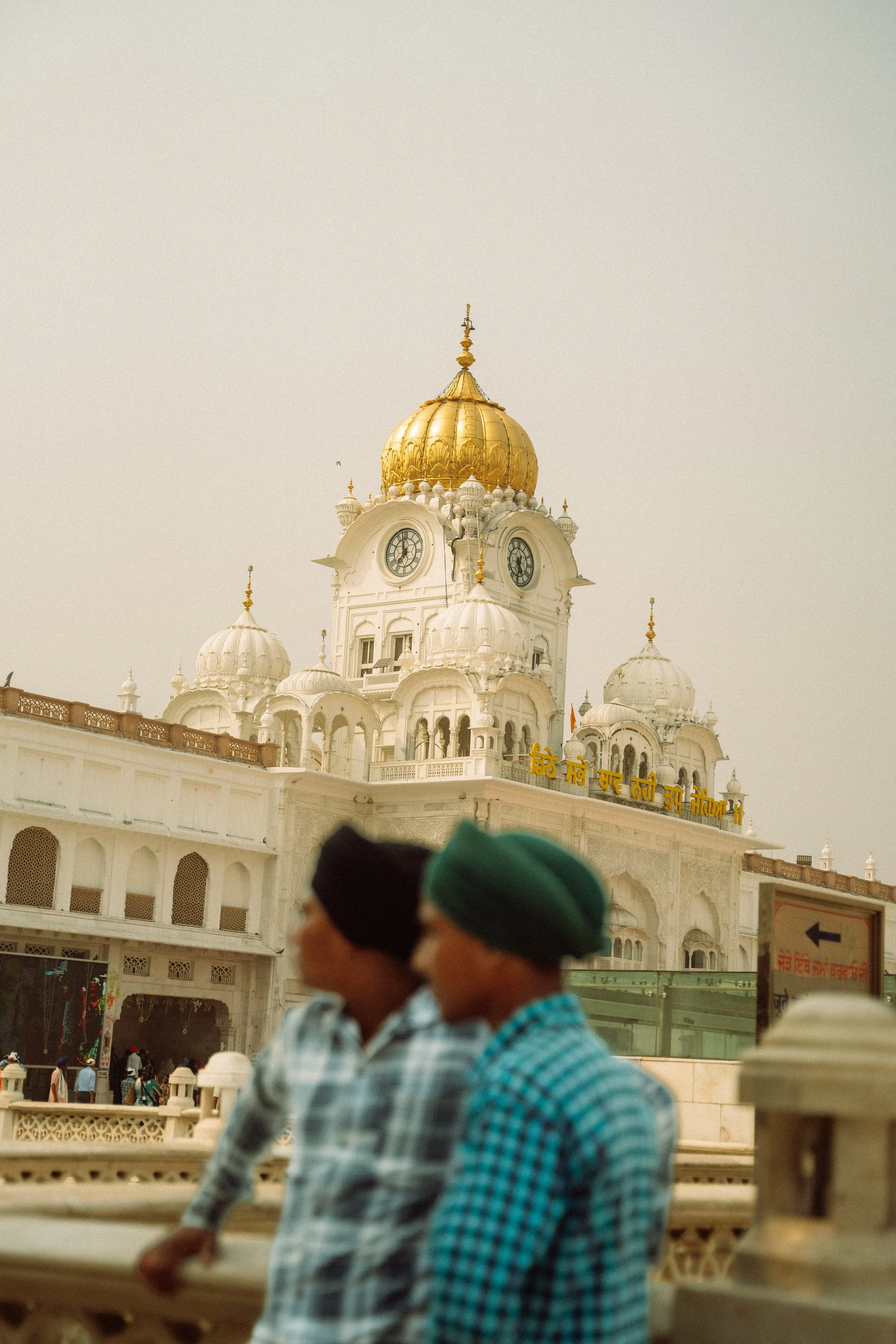 Blick auf das Goldene Tempel-Komplex in Amritsar, mit zwei Männern im Vordergrund, einer trägt einen schwarzen, der andere einen grünen Turban.