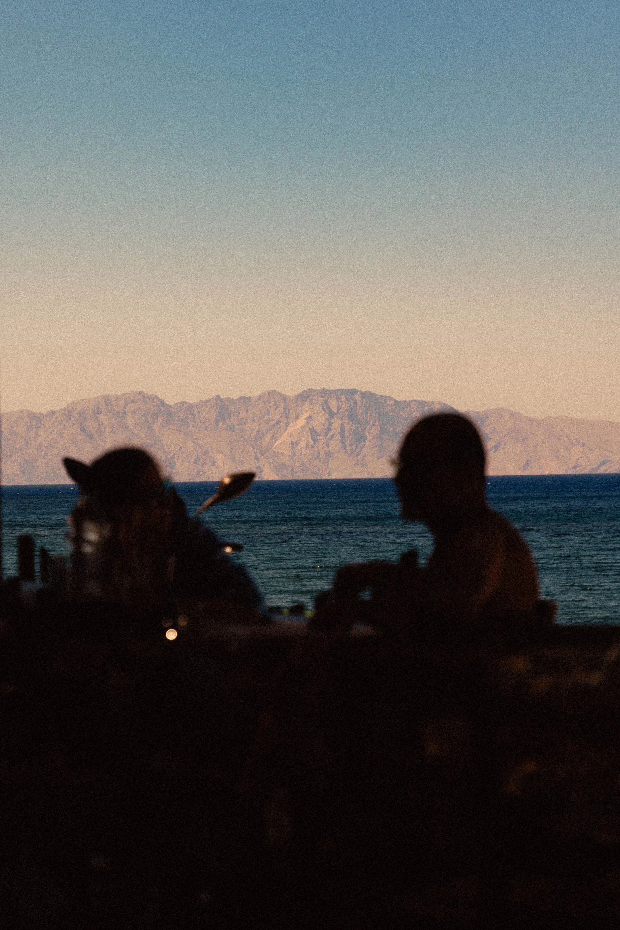 Silhouetten von zwei Personen, eine mit Pferdeohr, bei Sonnenuntergang am Strand mit Blick auf das Meer und Berge im Hintergrund.