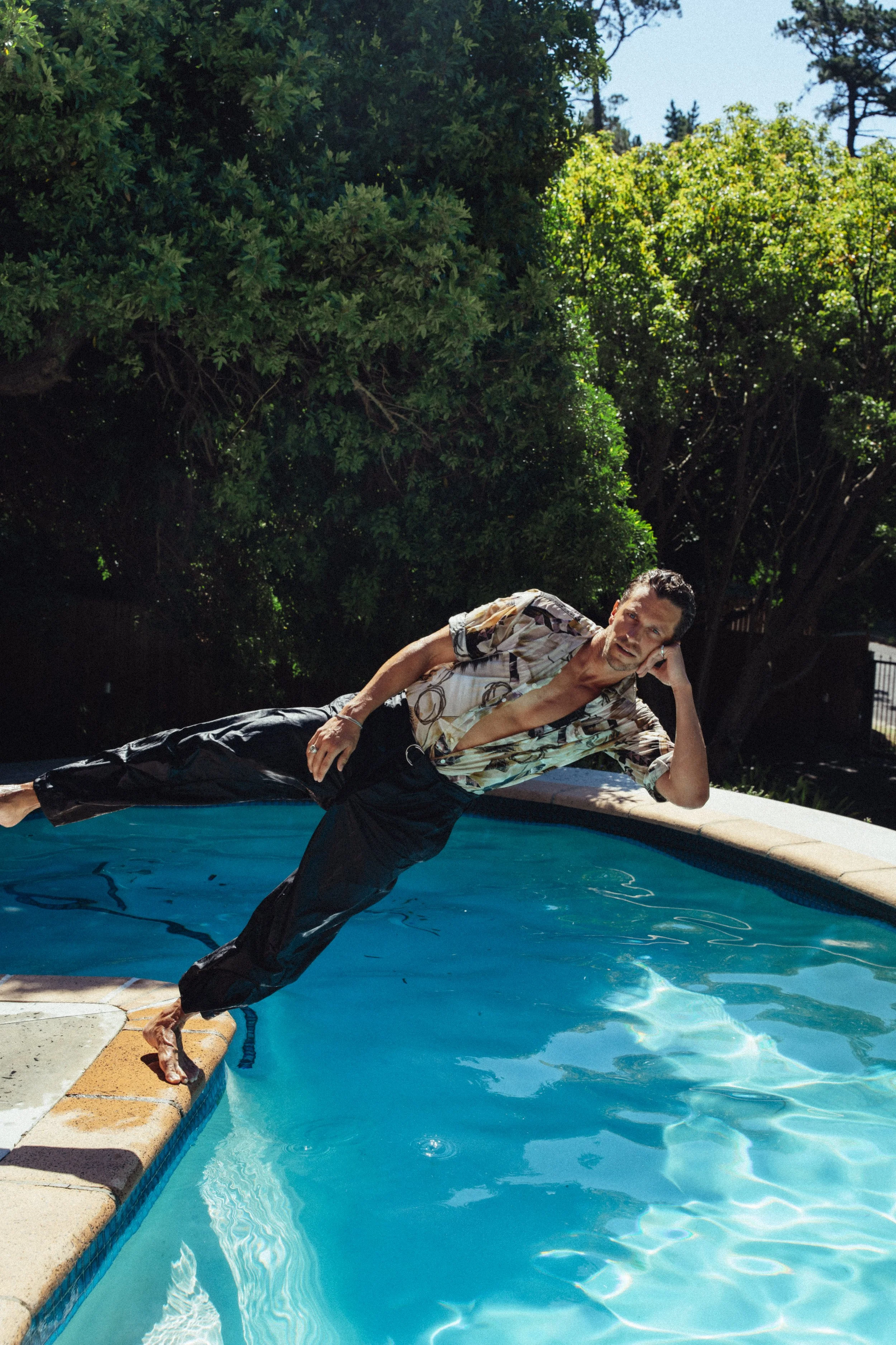 Mann, der an einer Poolkante lehnt, während er im Wasser liegt, bei sonnigem Wetter und grünen Bäumen im Hintergrund.