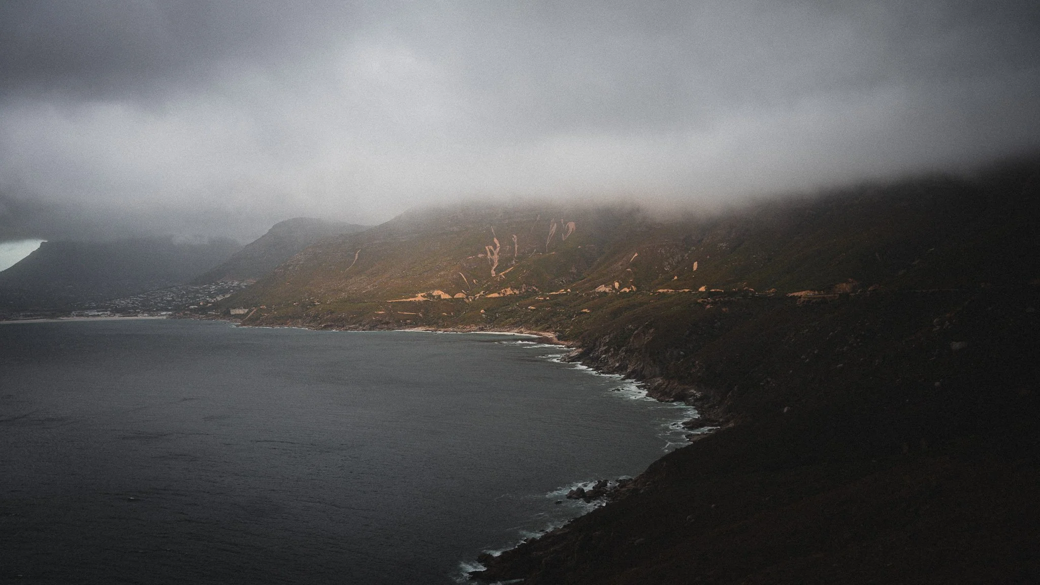 Küstenlandschaft mit dunklen Wolken, grauem Himmel, steilen Hügeln und dem Meer, das gegen die Klippen schlägt.