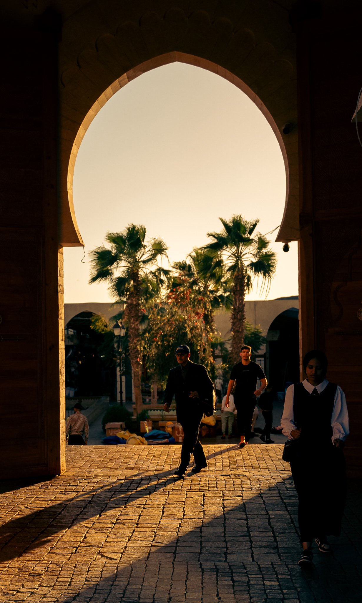 Menschen gehen durch ein Tor in der Abenddämmerung, Palmen im Hintergrund, lange Schatten auf gepflastertem Boden.
