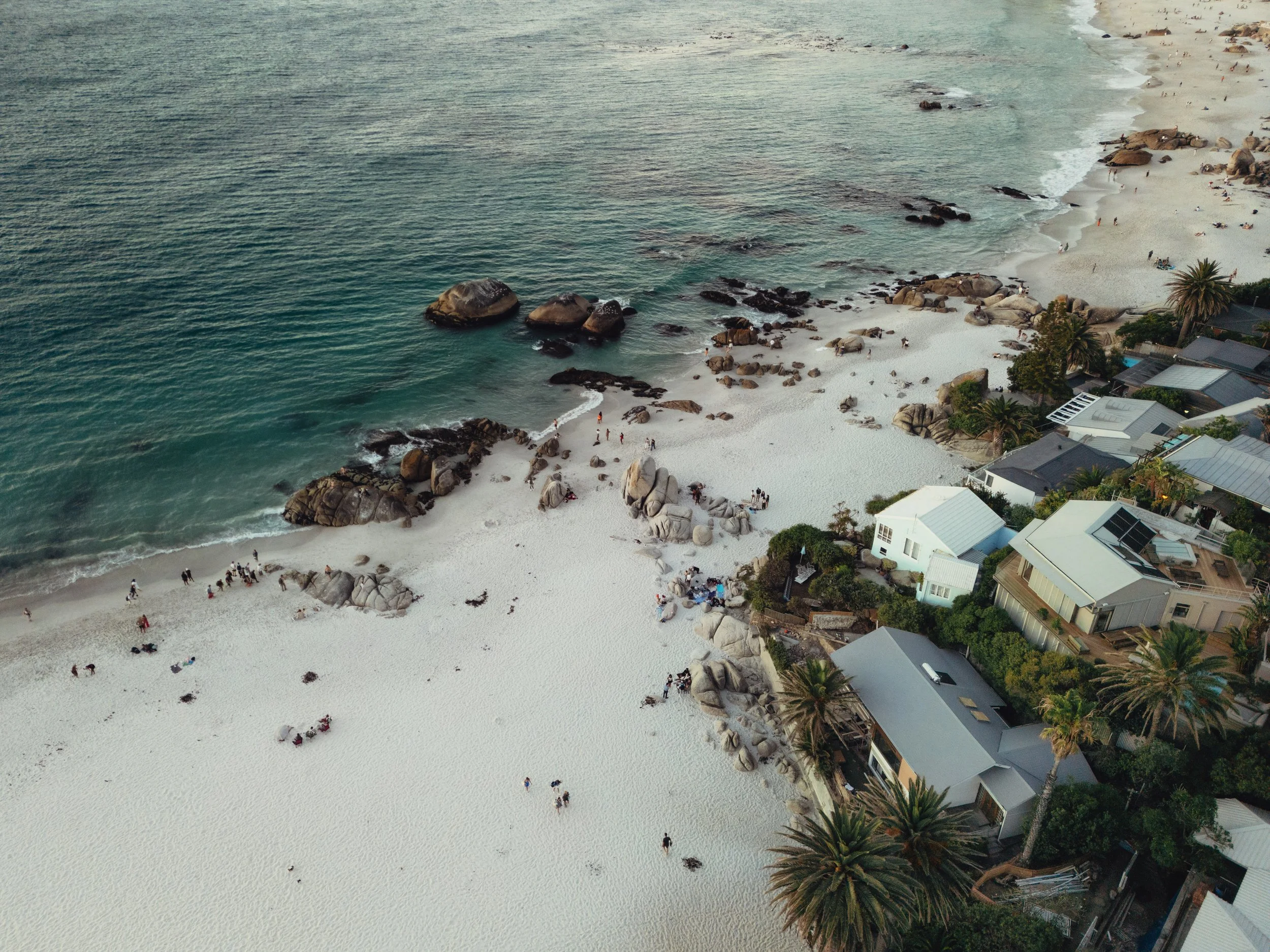 Luftaufnahme eines Strandes mit weißen Sand und großen Felsen, umgeben von Häusern und Palmen, mit mehreren Menschen im Sand und im Wasser.