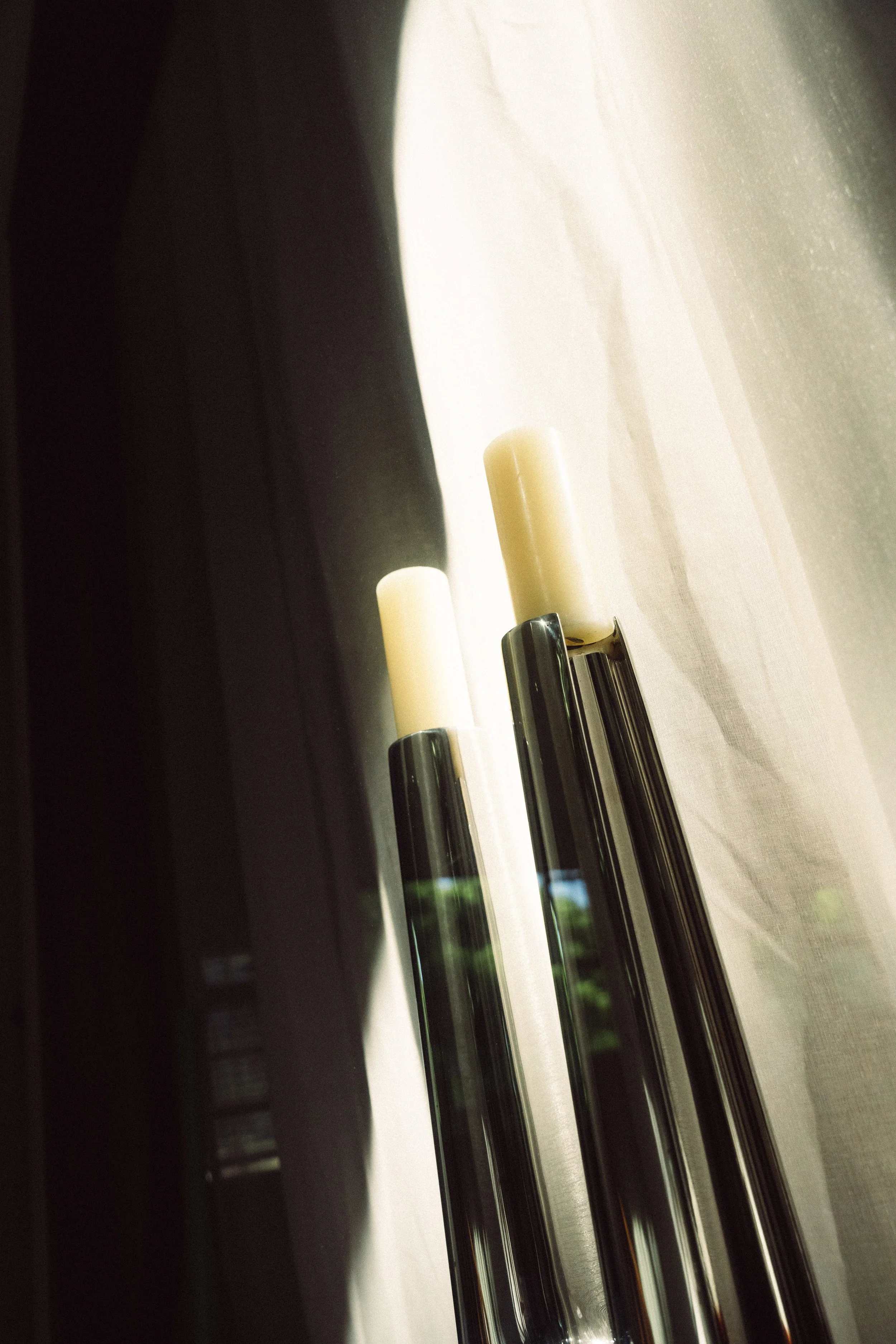 Zwei schwarze Kerzenhalter mit weißen Kerzen vor einem Fenster mit hellen Vorhängen.