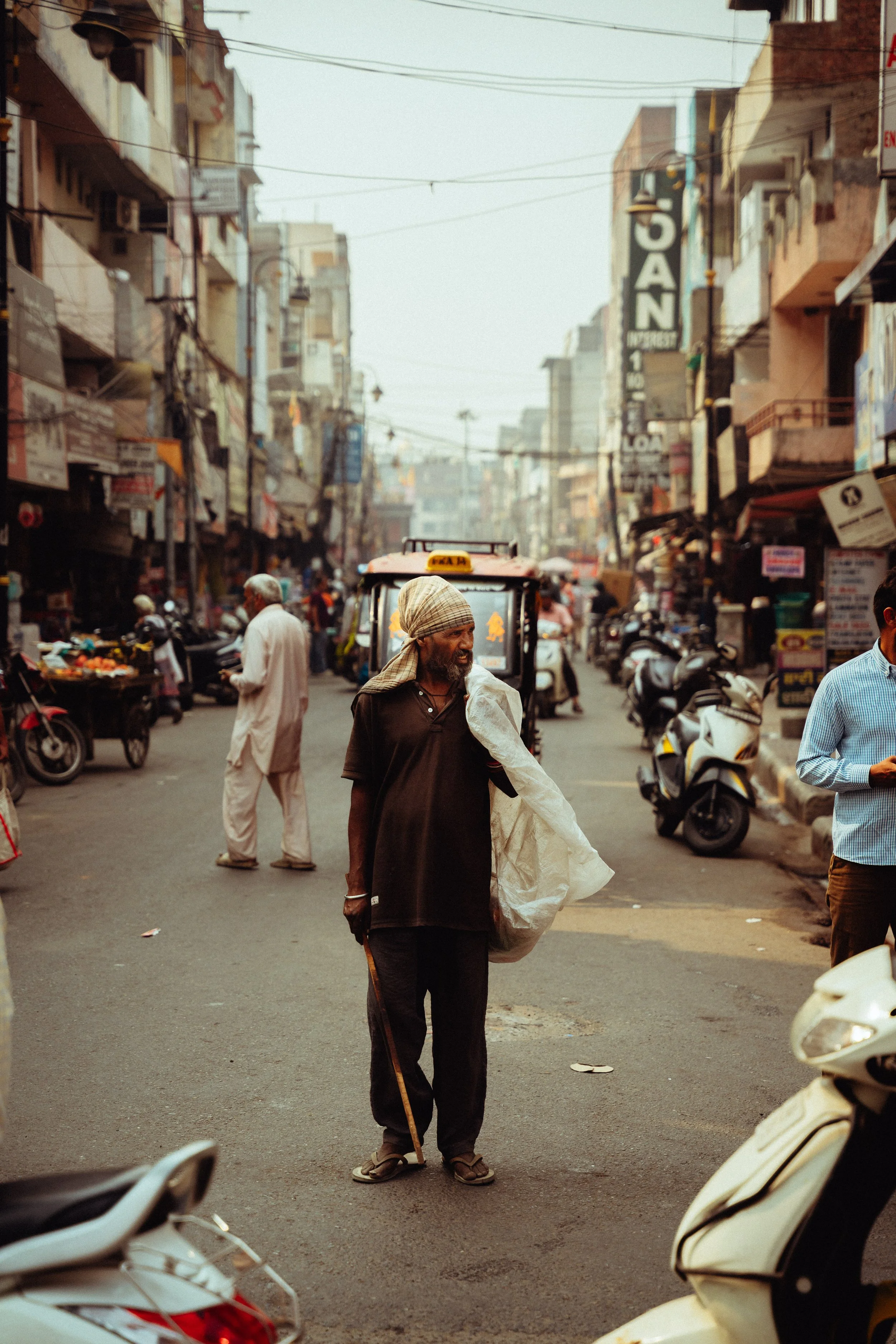 Straßen scene in einer belebten Stadt, ein älterer Mann mit Kopfhaube steht in der Mitte, umgeben von Leuten, Motorrädern und Geschäften auf beiden Seiten der Straße.