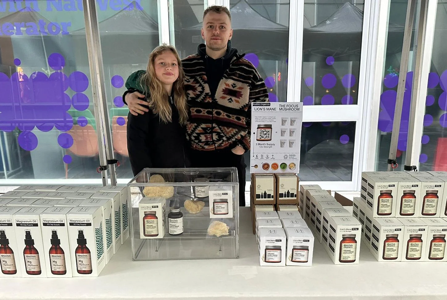 A man and a girl standing behind a table displaying various health supplement products, including boxes and bottles, in a bright indoor setting.