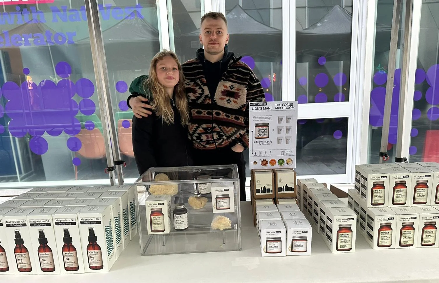 Two people, a man and a girl, standing behind a table displaying various health supplement products, including boxes of magnesium oil and bottles of mushroom extract, at an outdoor market or event.