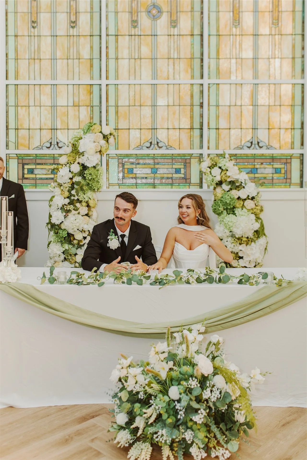Bride and groom seated together at sweetheart table beneath stained glass windows
