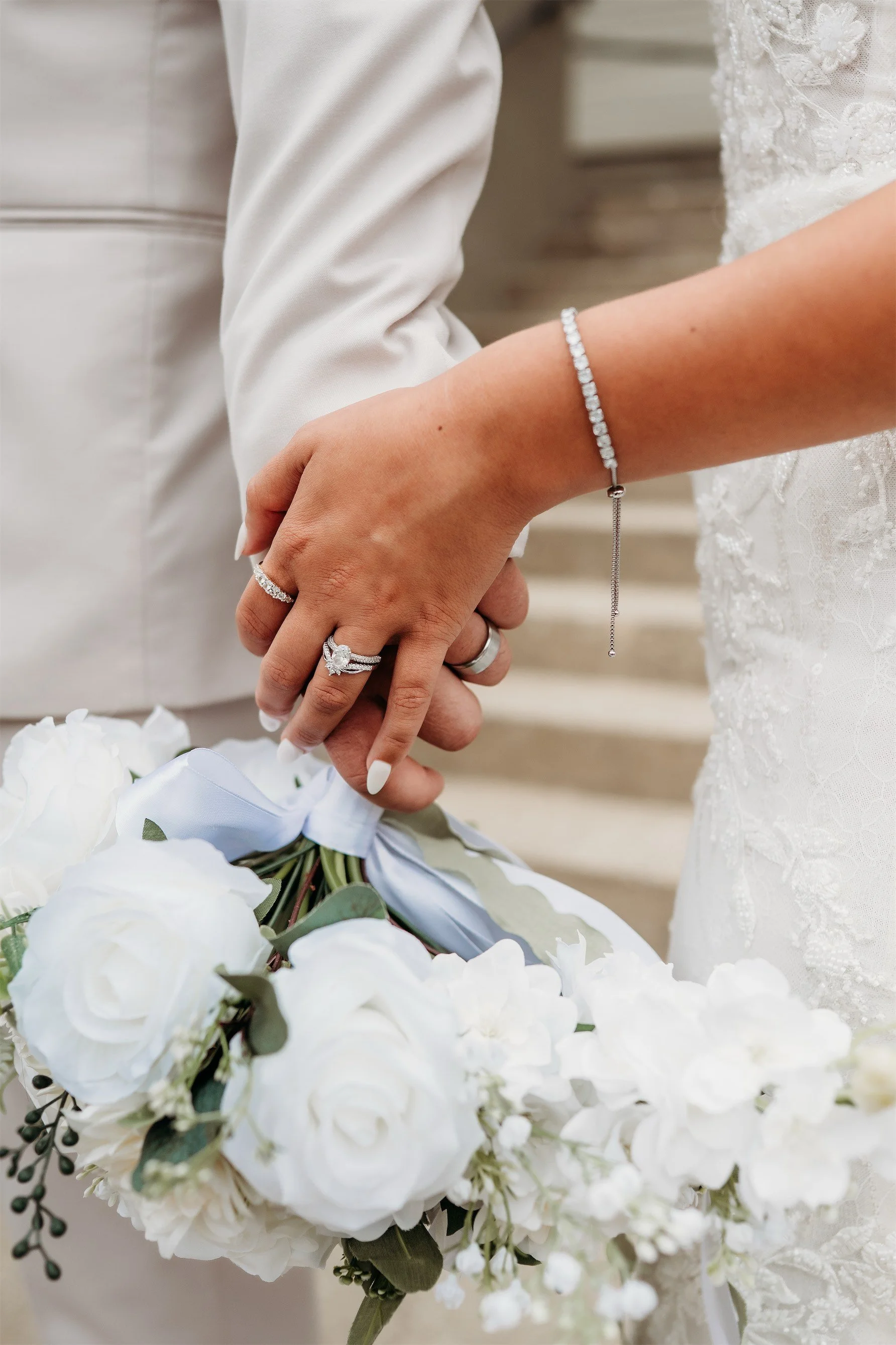 Close up of the couple's clasped hands showing diamond rings beside a white rose bouquet with blue ribbon