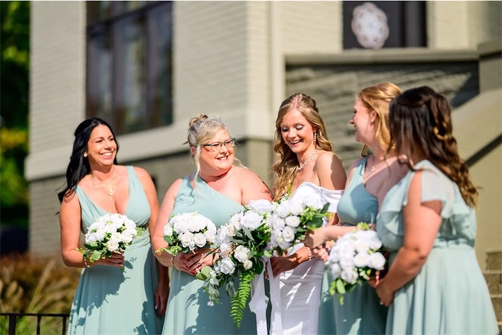 Bridesmaids in dusty sage dresses hold white bouquets and laugh with the bride outside the church
