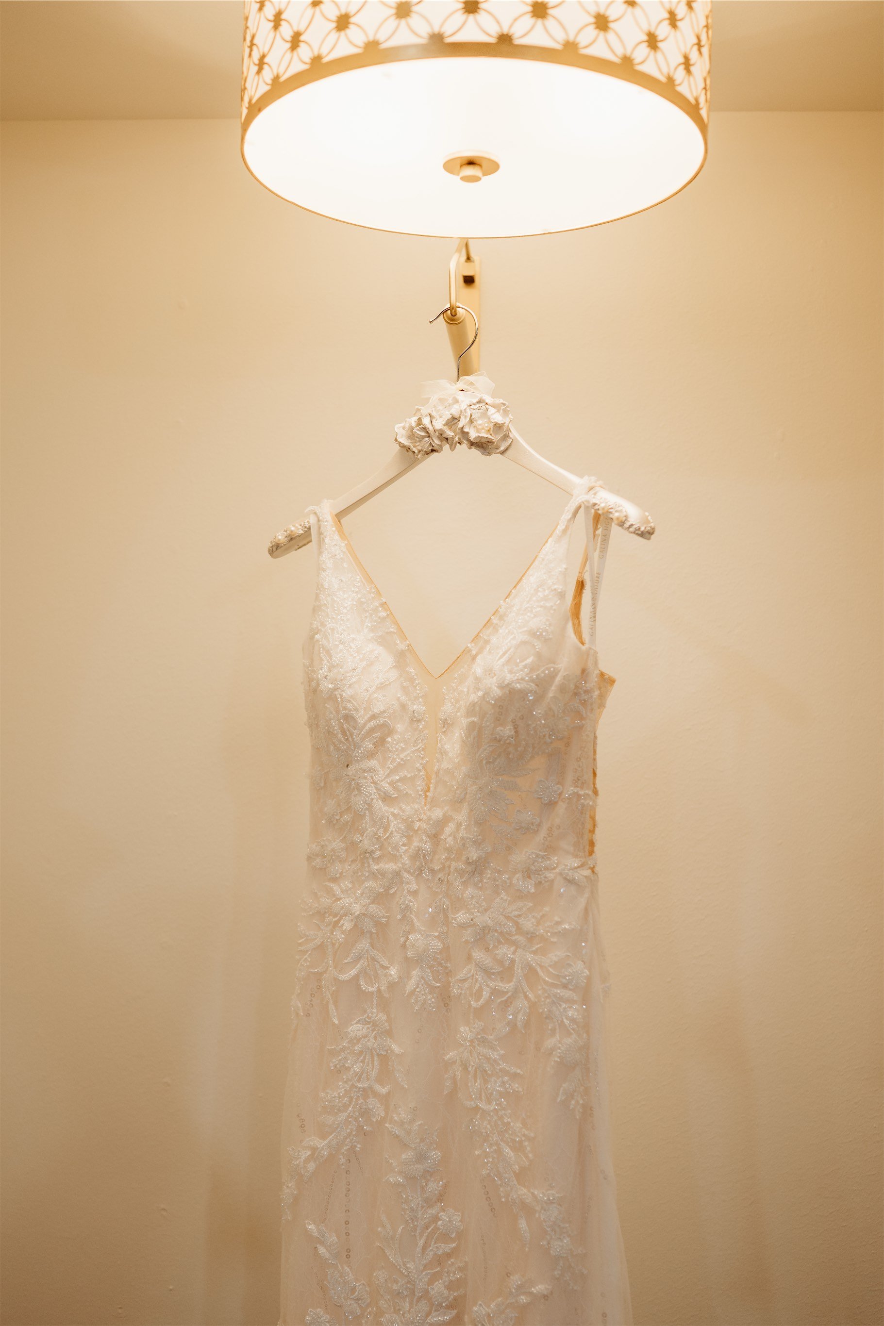 Lace wedding gown with a plunging V neckline hanging from a floral hanger beneath a warm glowing light