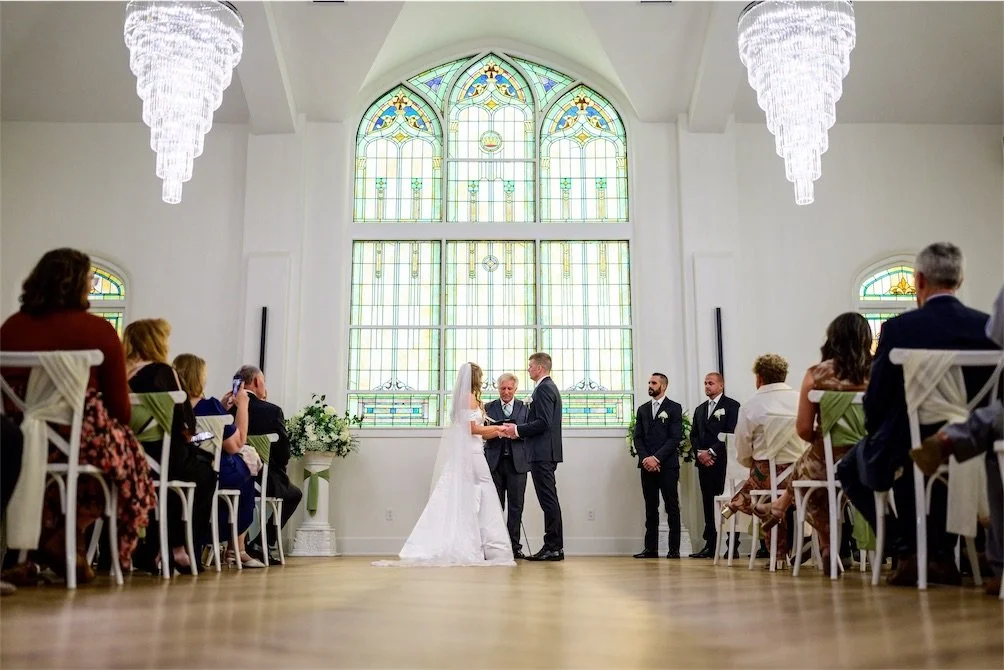 Couple exchanging vows at the altar while guests sit on white chairs facing a stained glass window
