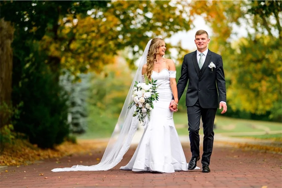 Bride in a long veil and groom in a charcoal suit hold hands on a brick path under autumn trees