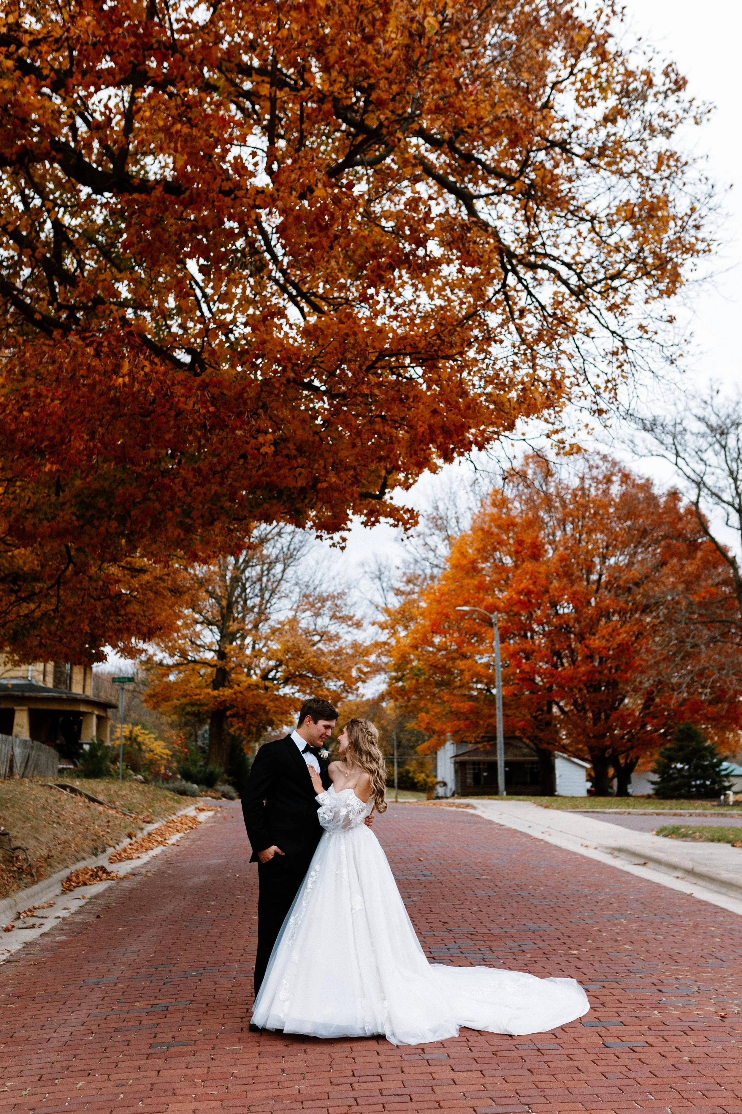 A bride and groom standing on a brick street surrounded by fall trees with orange and red leaves, in a quiet neighborhood.