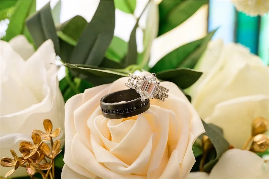 Diamond engagement ring and black wedding band resting on an ivory rose among greenery