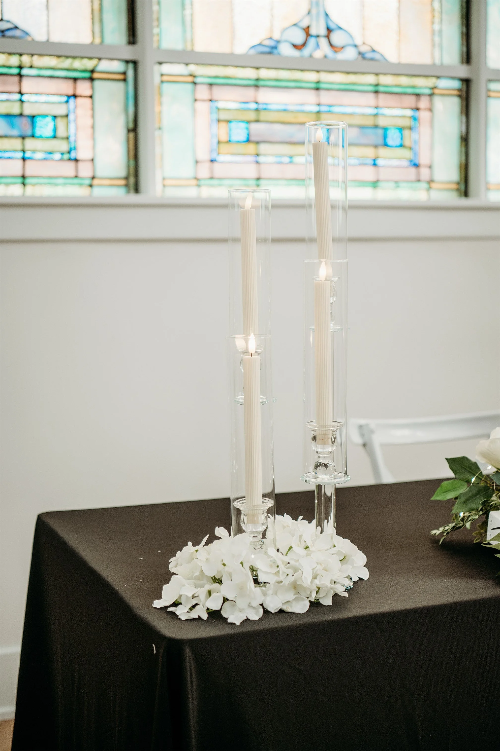 Tall glass hurricane candle holders with white taper candles and white flower petals on a black draped table