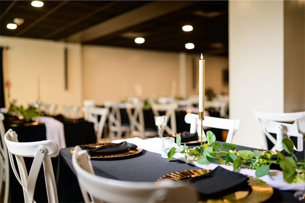 Reception table set with black linens, gold chargers, a taper candle and greenery garland beside white chairs
