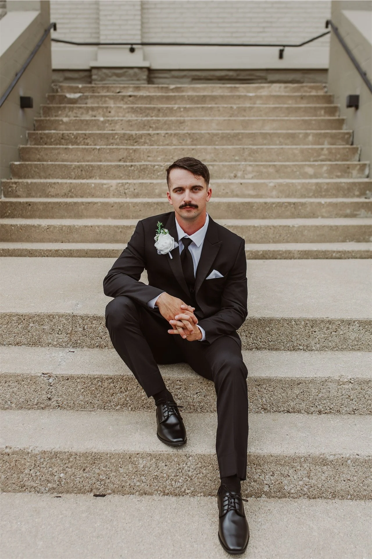 Groom in black suit sitting on stone steps outside church wedding venue