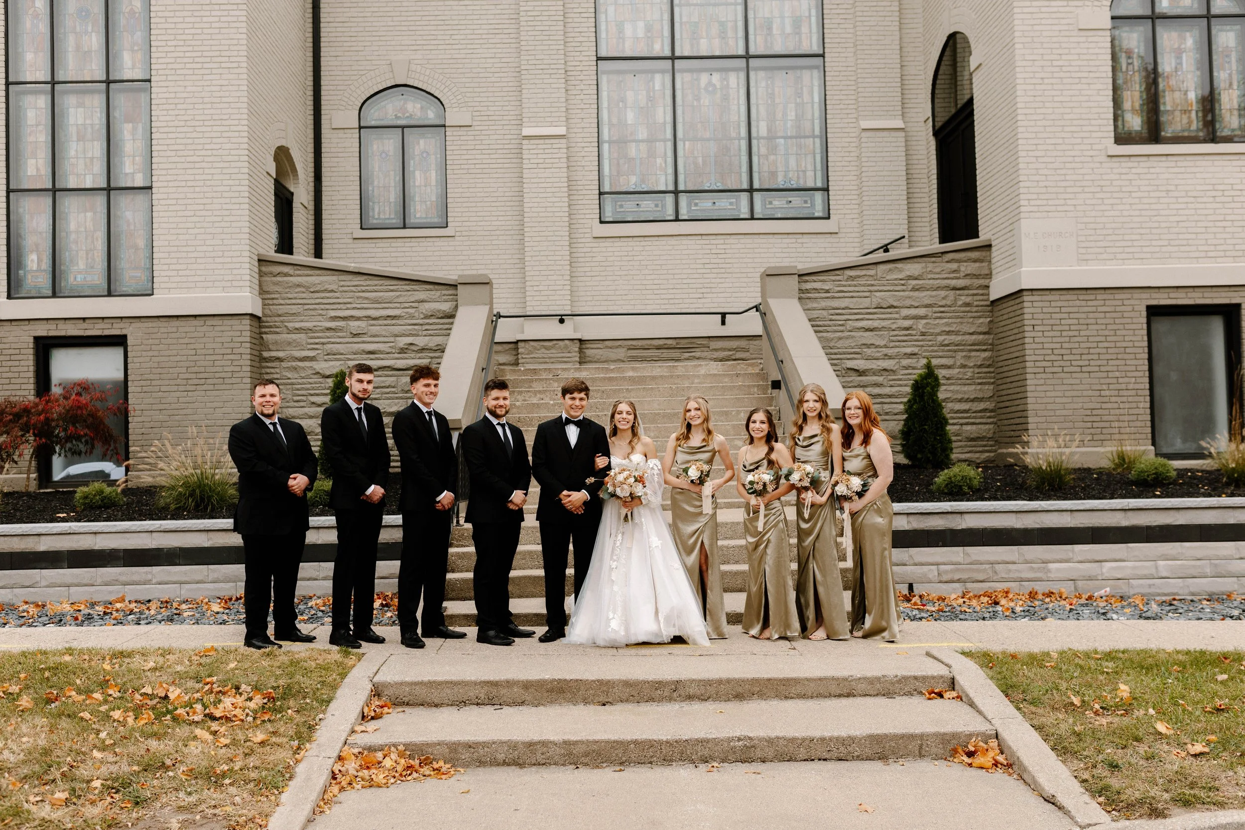 A group of eight people in formal wedding attire standing on steps outside a brick building with large windows.