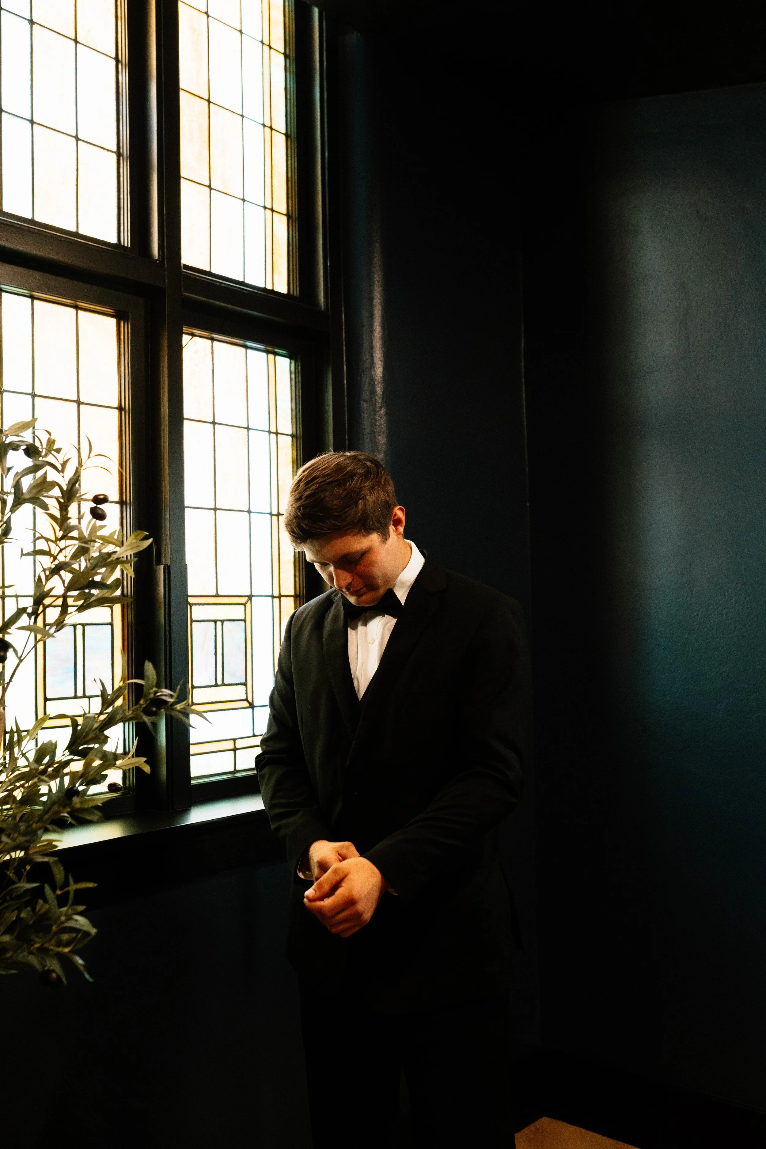 A young man in a tuxedo standing indoors near a large stained glass window, looking down with his hands clasped.